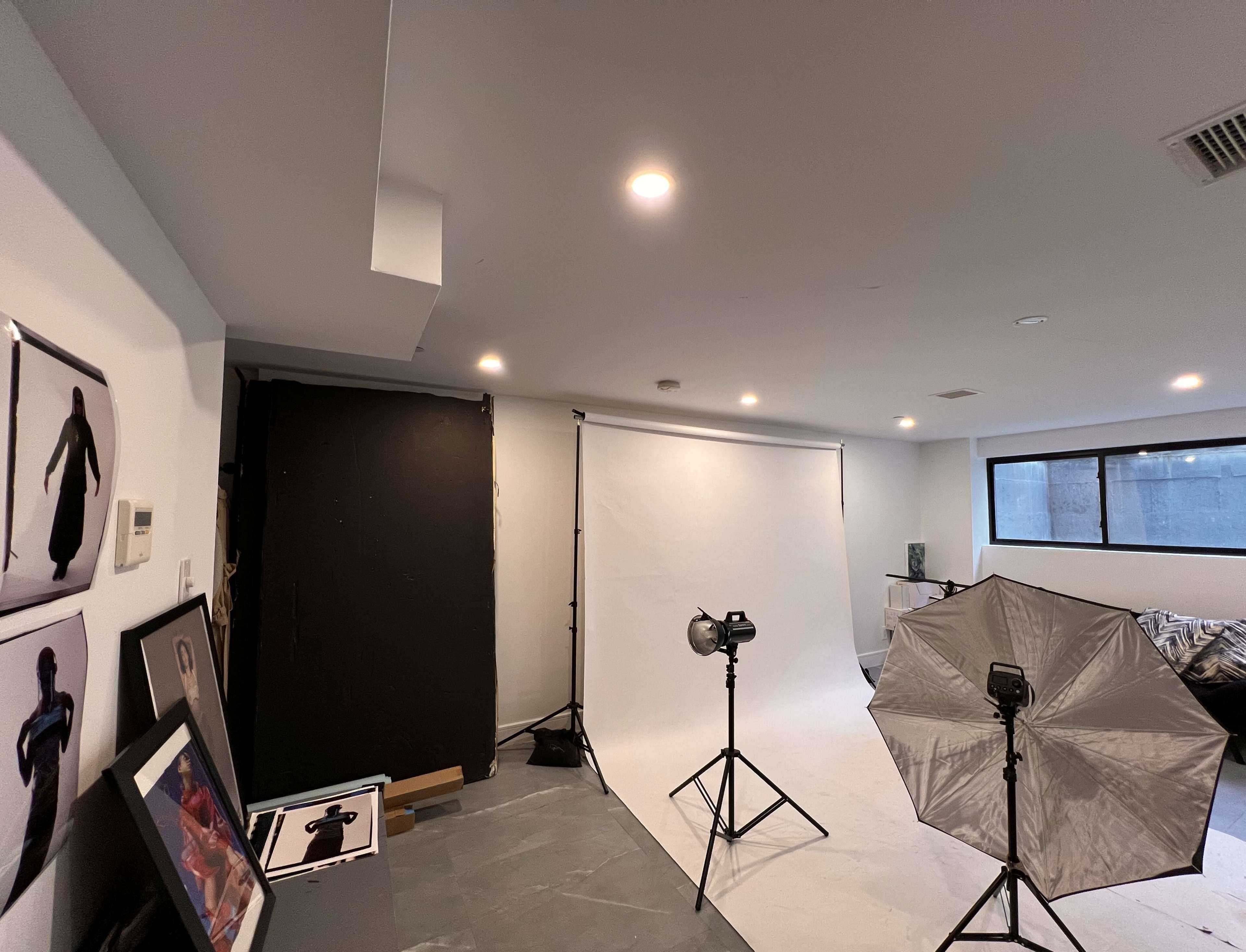 The image shows a photography studio setup with a white backdrop, lighting equipment, and various framed photographs on a wall.