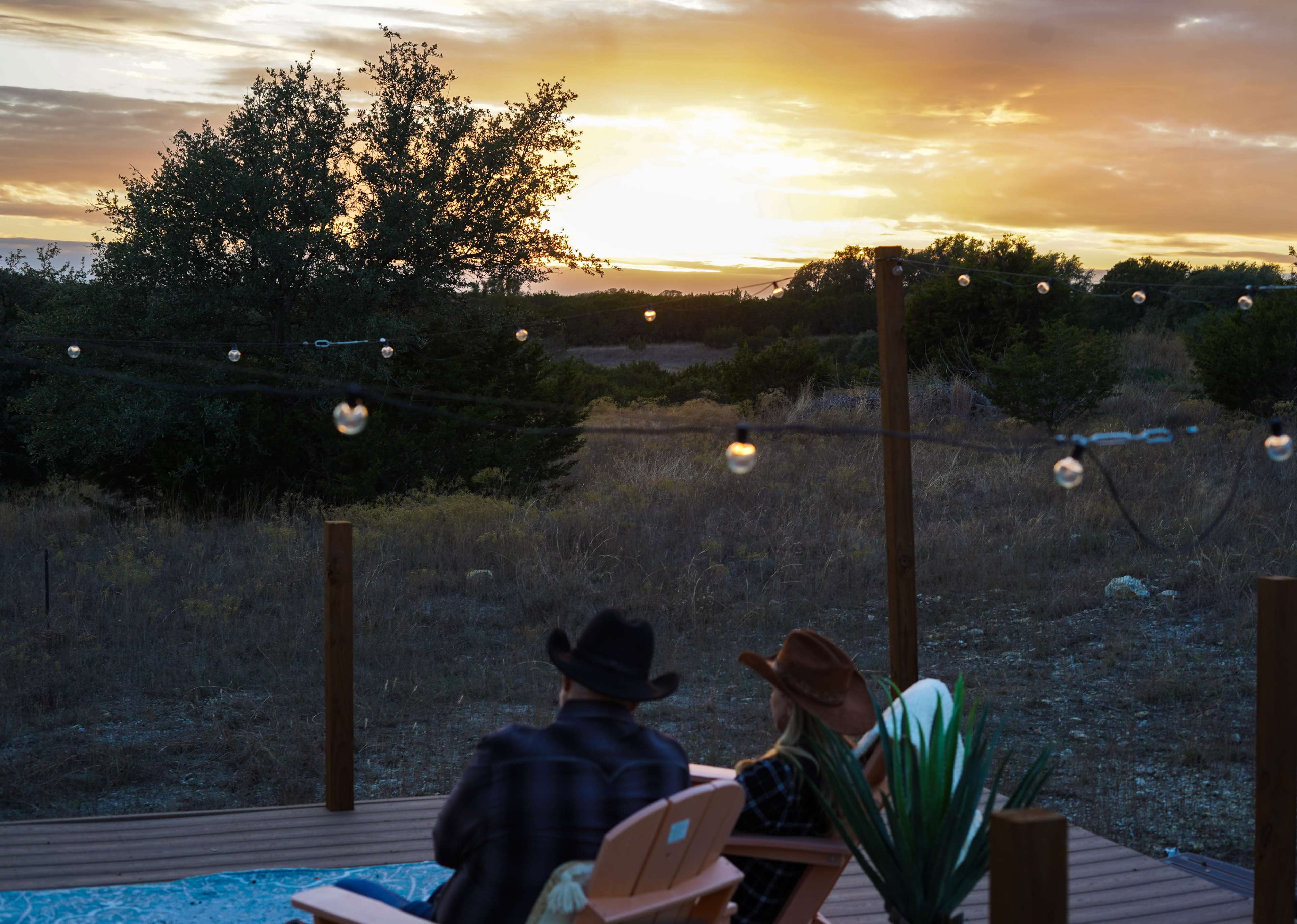 Two people in cowboy hats sit on a deck overlooking a sunset with string lights hanging nearby.