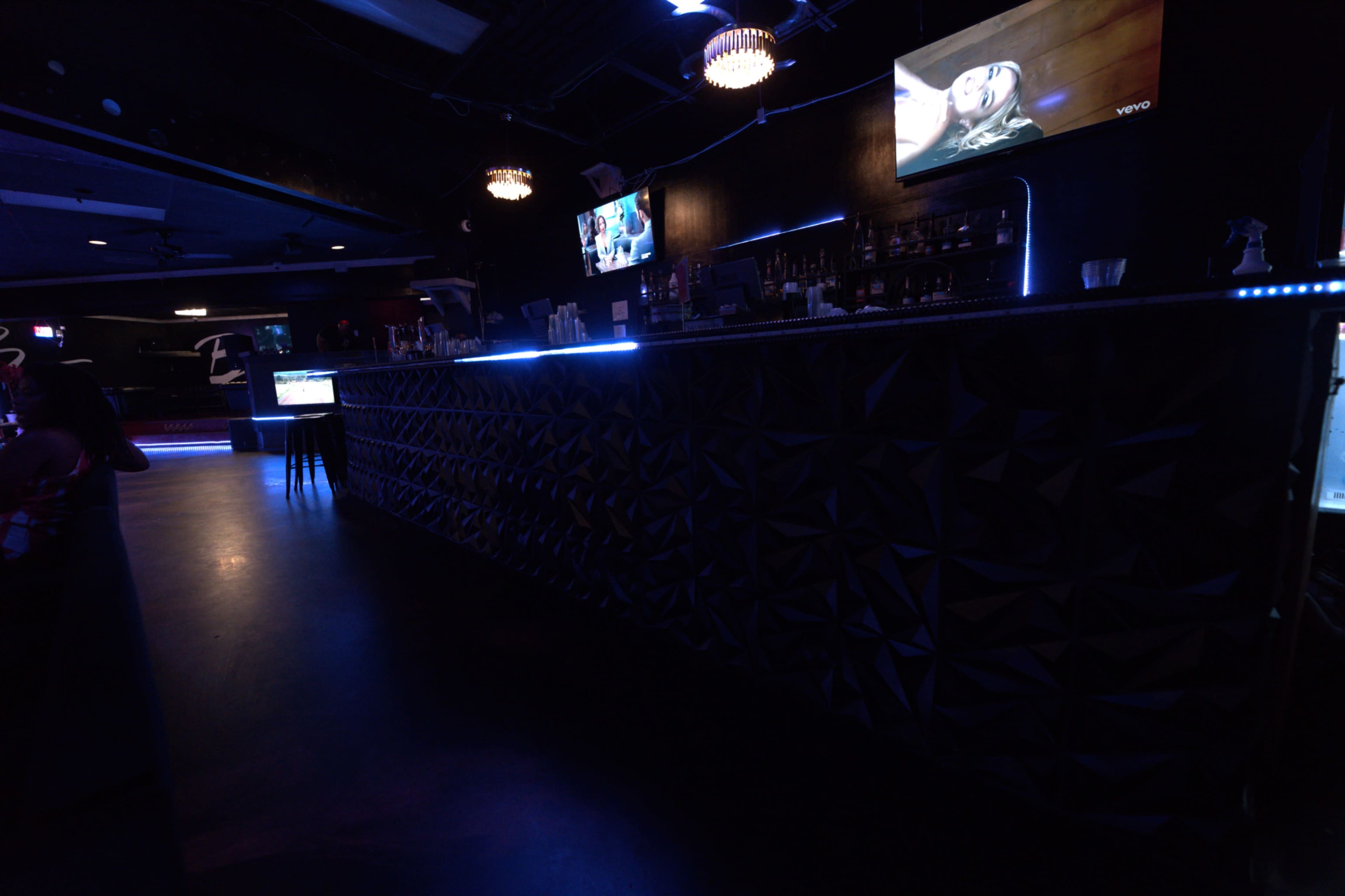 The image shows a dimly lit bar with a textured black counter, illuminated by blue LED lights, and two televisions mounted on the wall.
