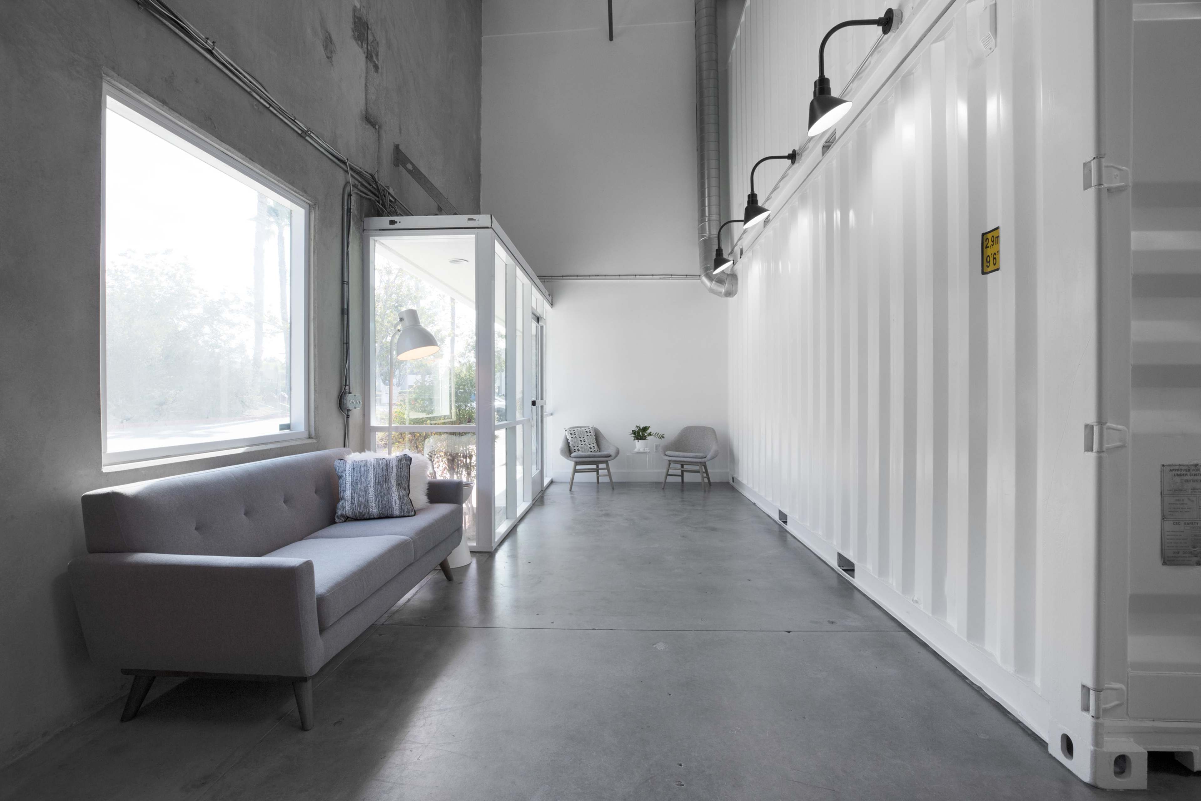 A modern interior space featuring a gray sofa, two chairs, and large windows, with a corrugated shipping container on one side.