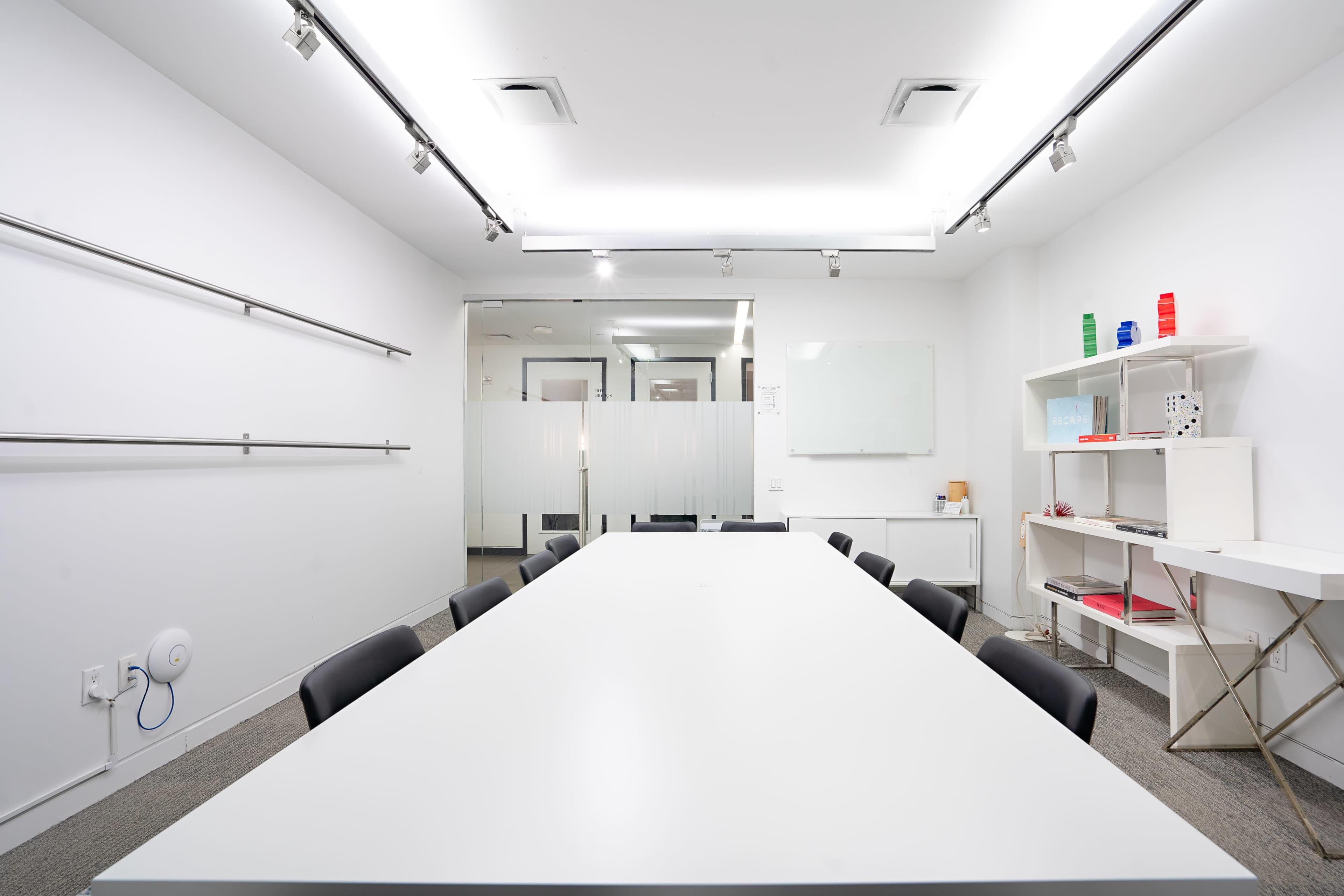 A large, empty conference room features a long white table surrounded by black chairs, with whiteboard and glass partitions in the background.