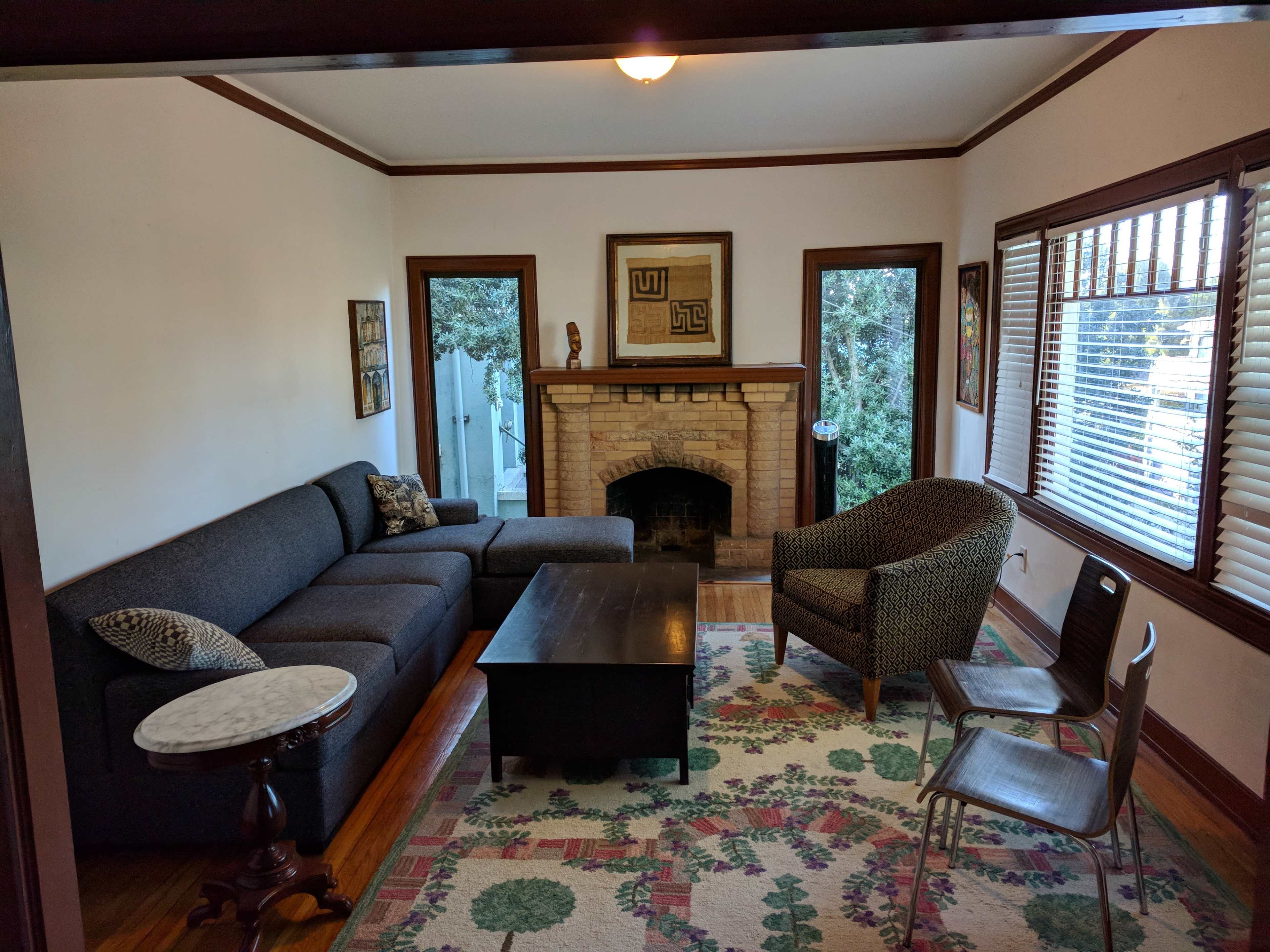 A living room with a gray sectional sofa, a dark coffee table, and a patterned rug, featuring a fireplace and multiple windows with blinds.