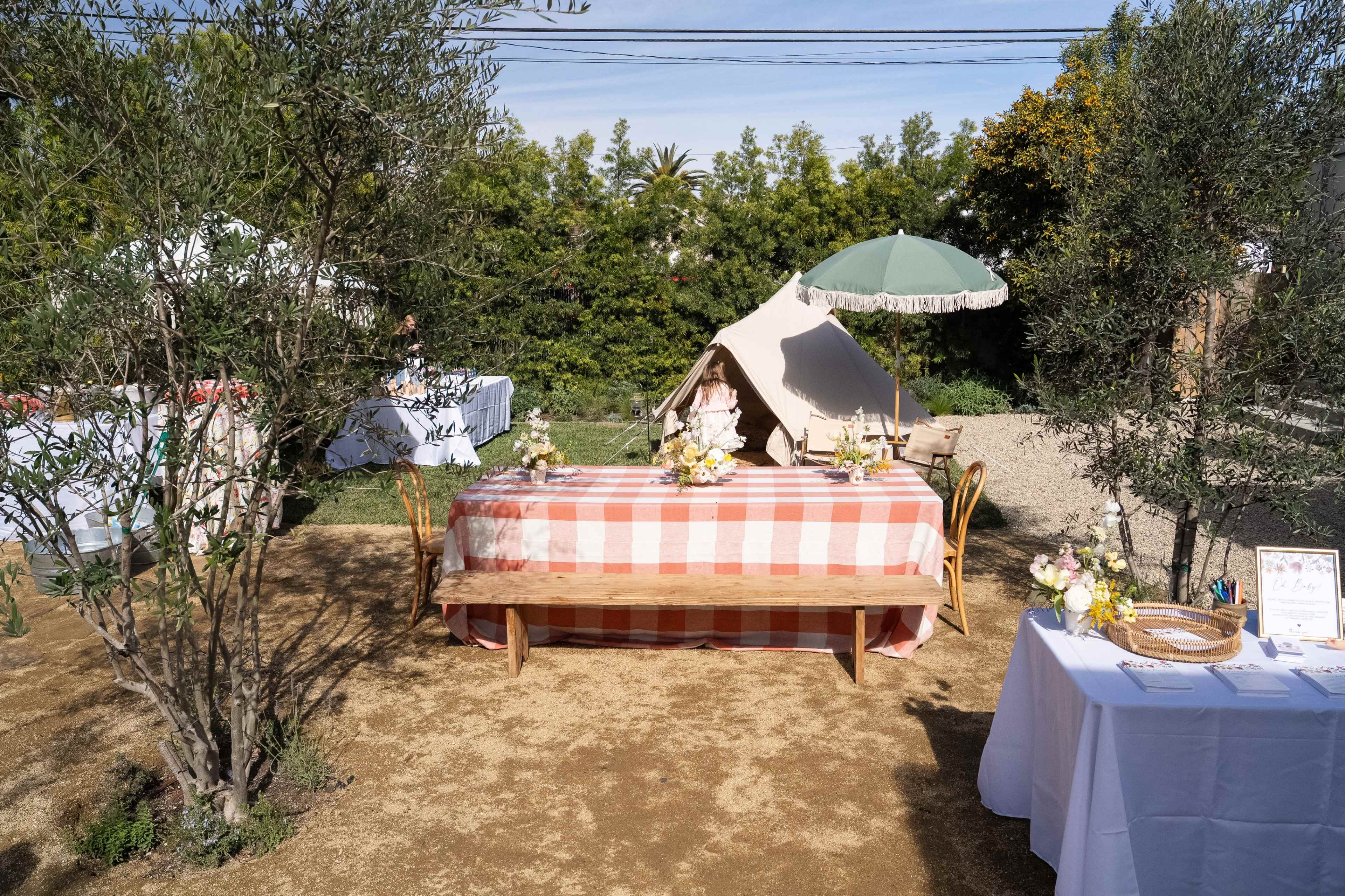 The scene features a picnic setup with a checkered tablecloth, decorated with flowers, surrounded by olive trees and a tent in an outdoor garden.