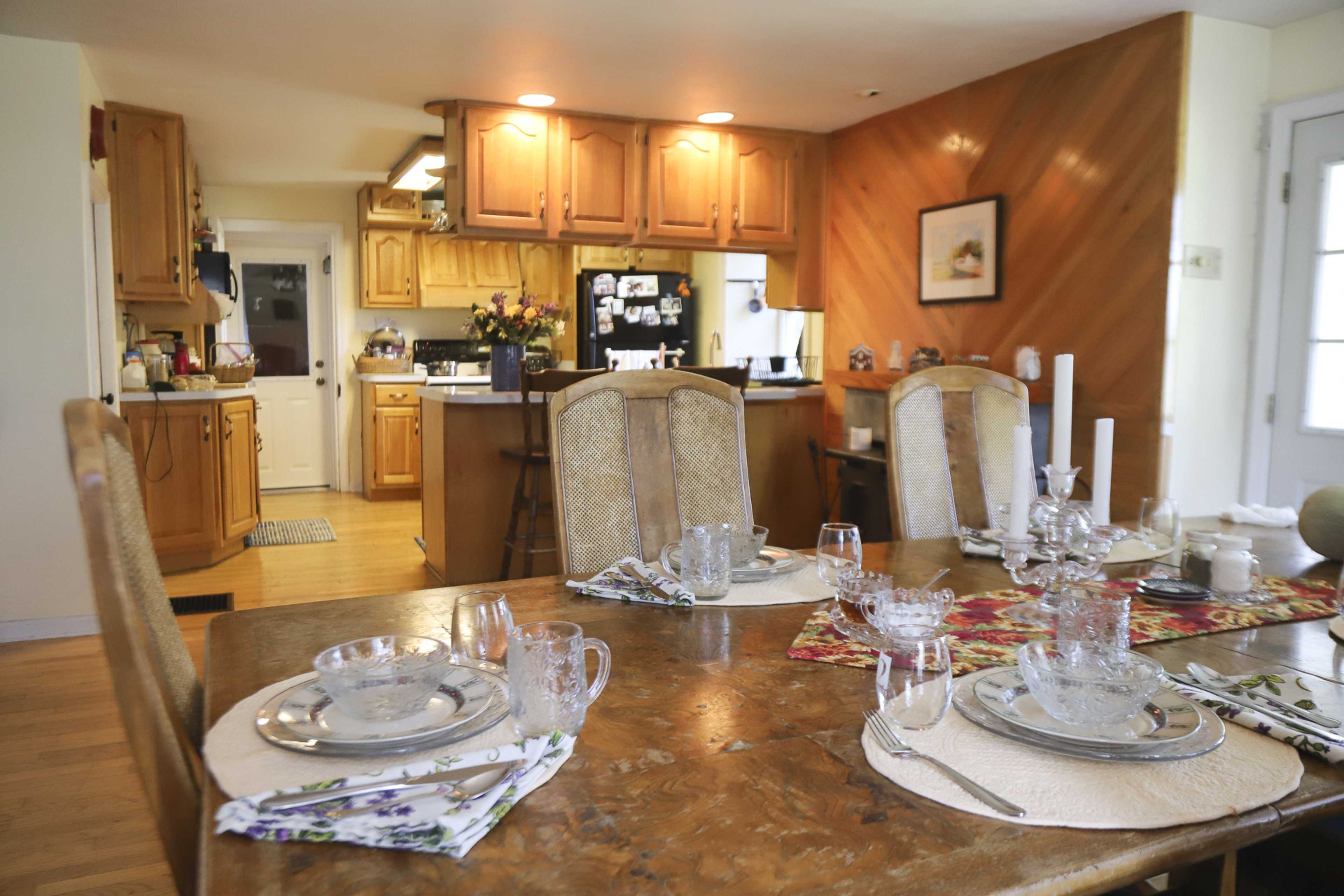 A dining table is set with glassware and place settings, with an open kitchen visible in the background.