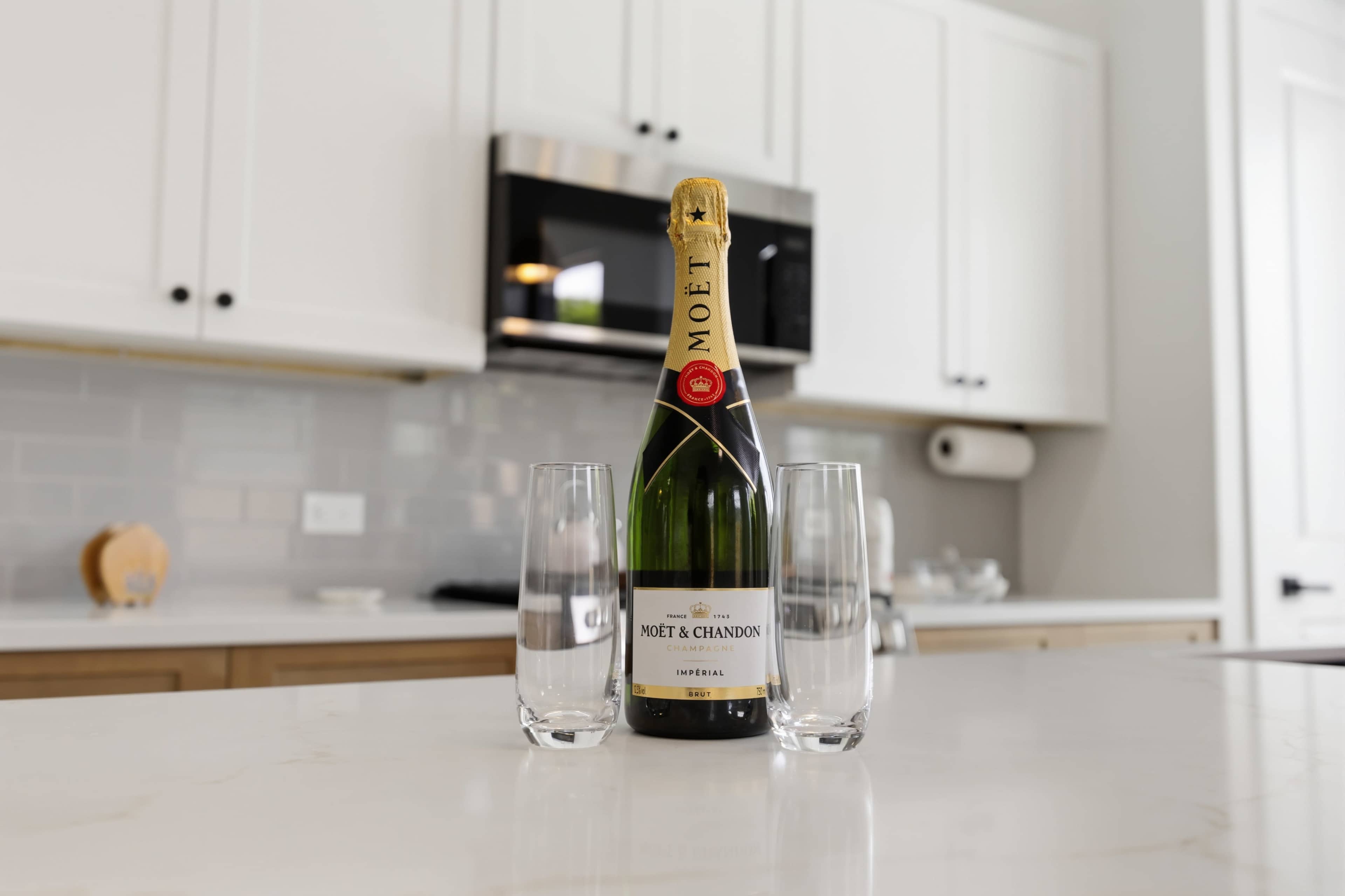 A bottle of Moët & Chandon champagne stands between two clear glass flutes on a white countertop in a modern kitchen.
