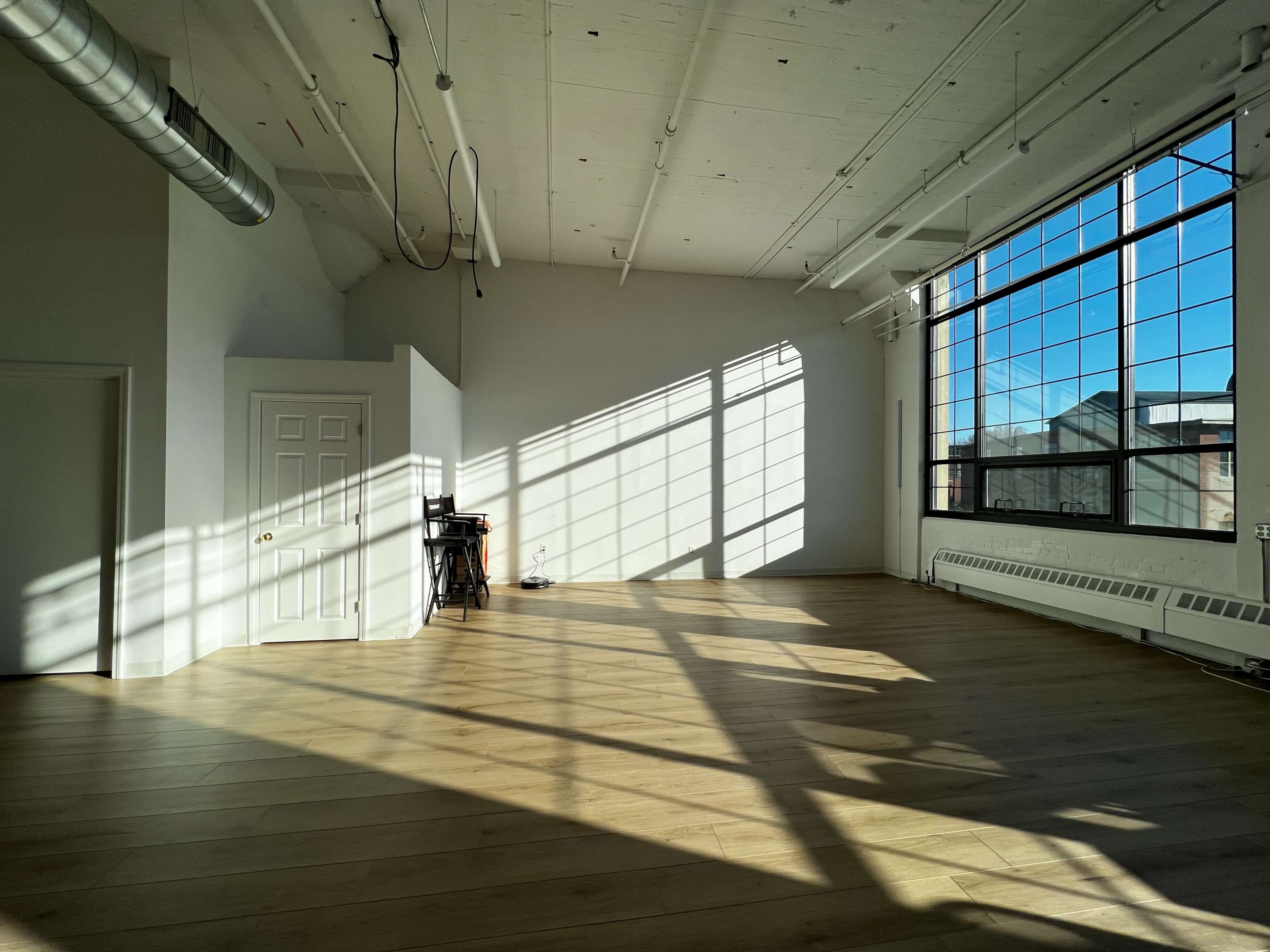 A spacious, sunlit room with wooden floors and large windows casting shadows across the walls.