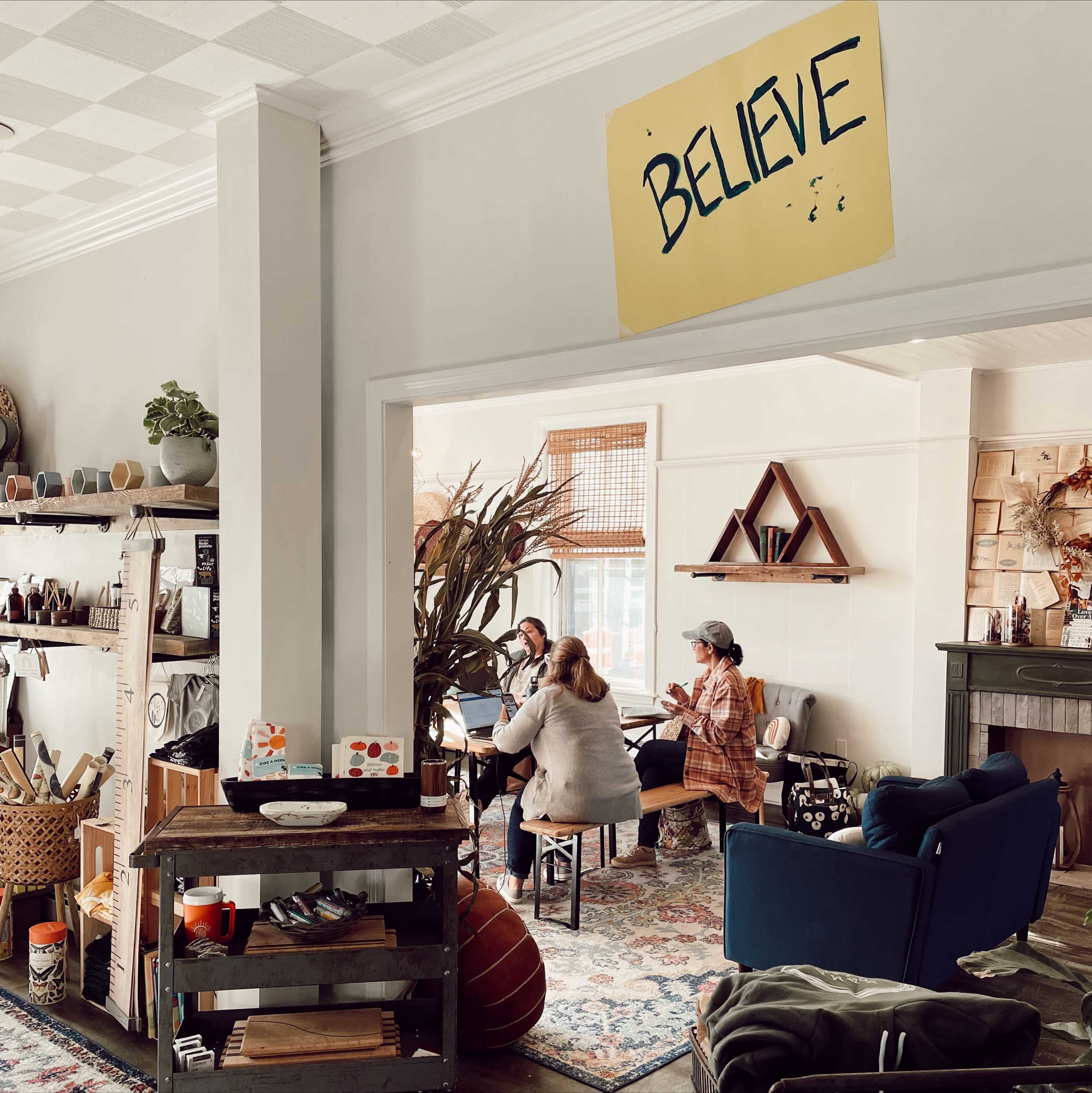 A cozy room features a group of three people interacting around a table, with various decorative items and plants visible throughout the space.