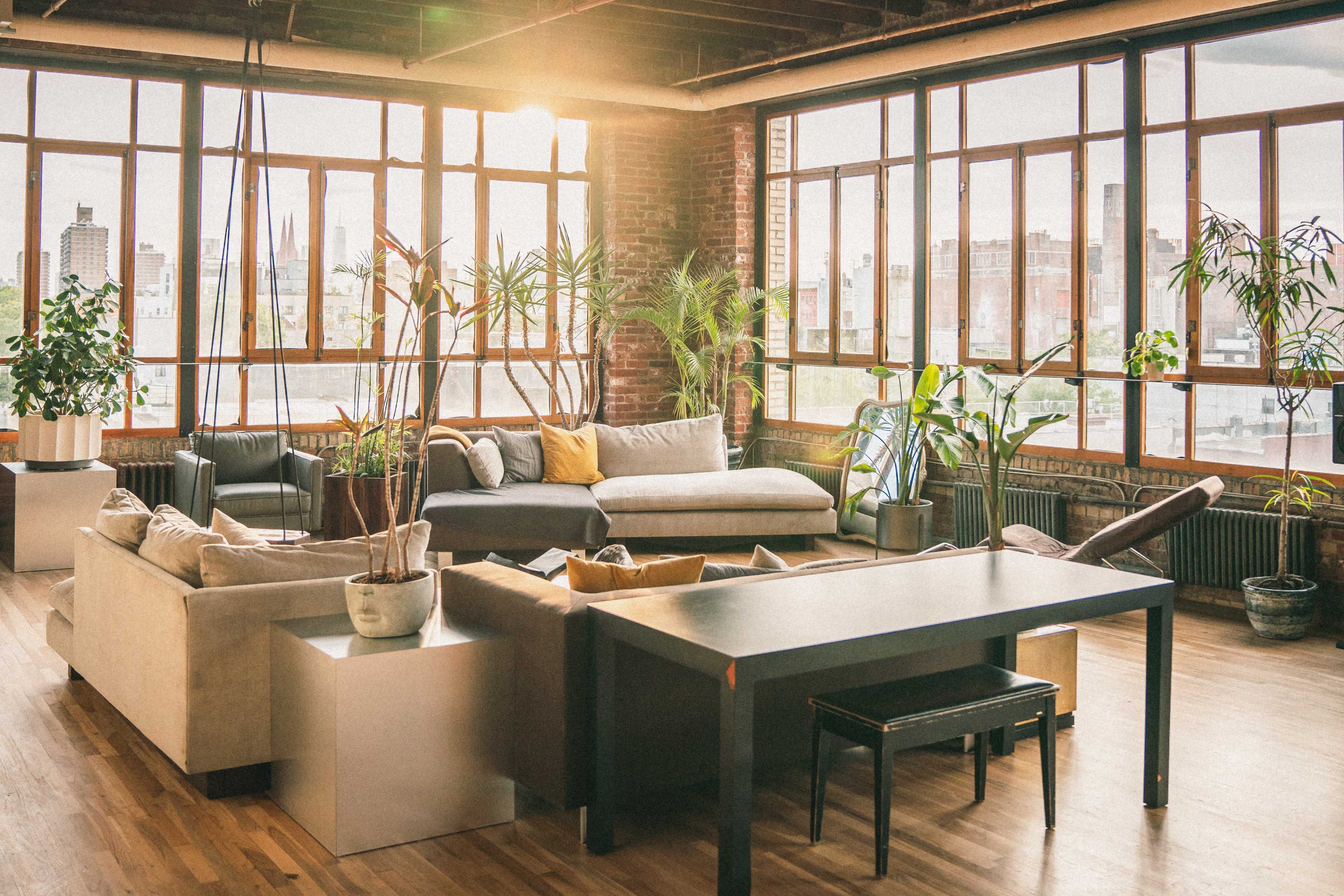 A spacious, industrial-style living room features large windows, plants, a comfortable sofa, and a dining table.