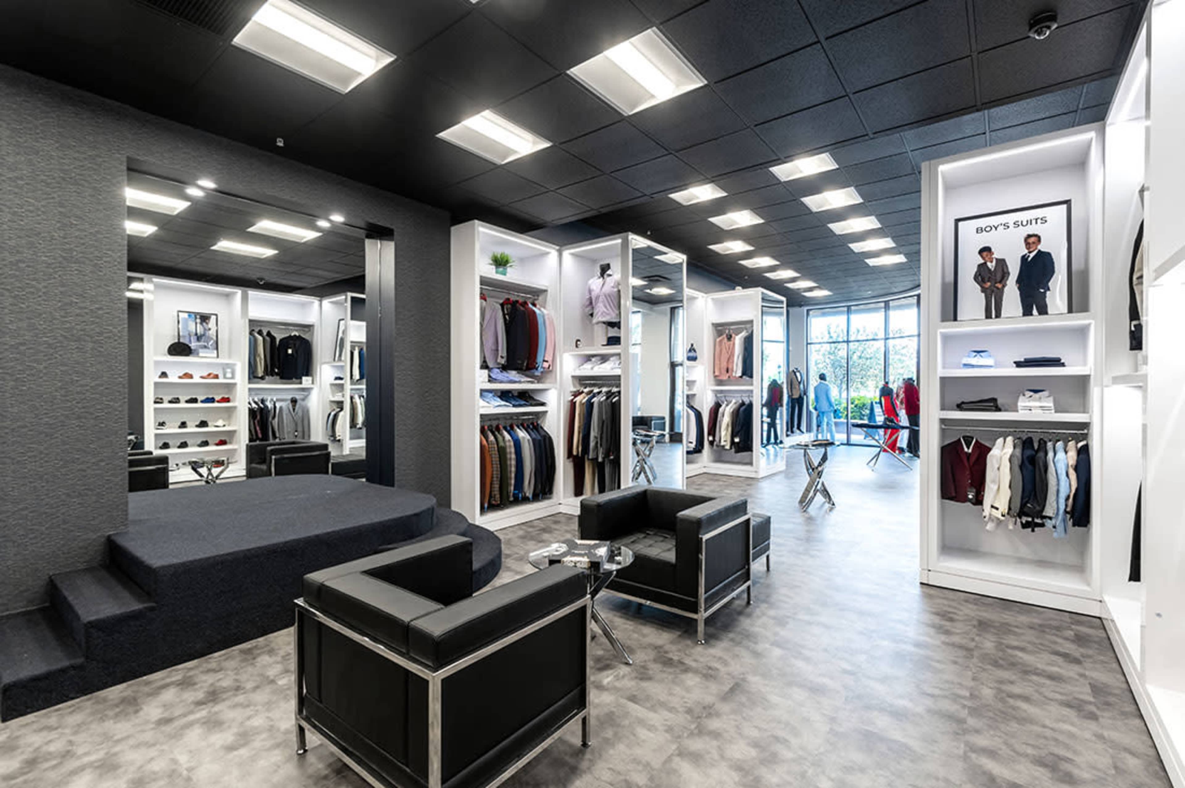 A modern retail clothing store featuring white display units with various garments, a seating area with black chairs, and large windows allowing natural light.
