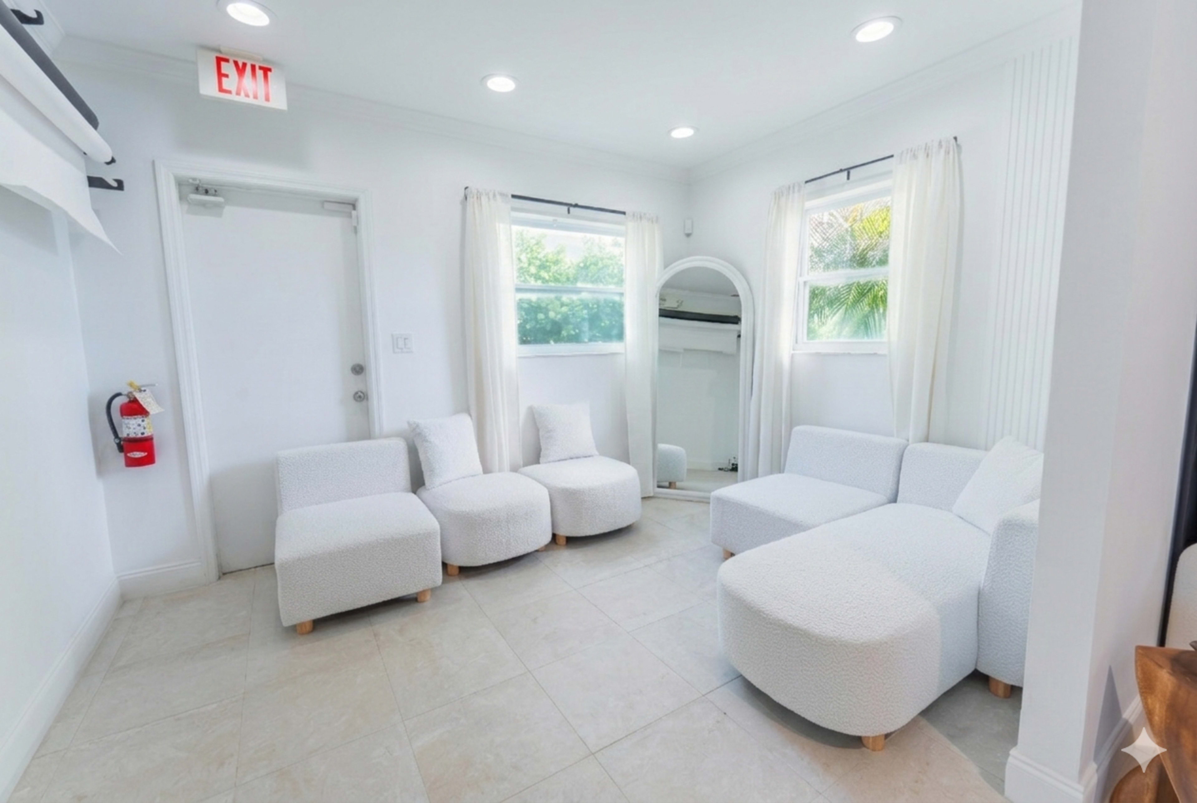 The room features a light-colored interior with a seating area comprised of three round chairs and a sectional sofa, along with a wall-mounted mirror and a door leading outside.