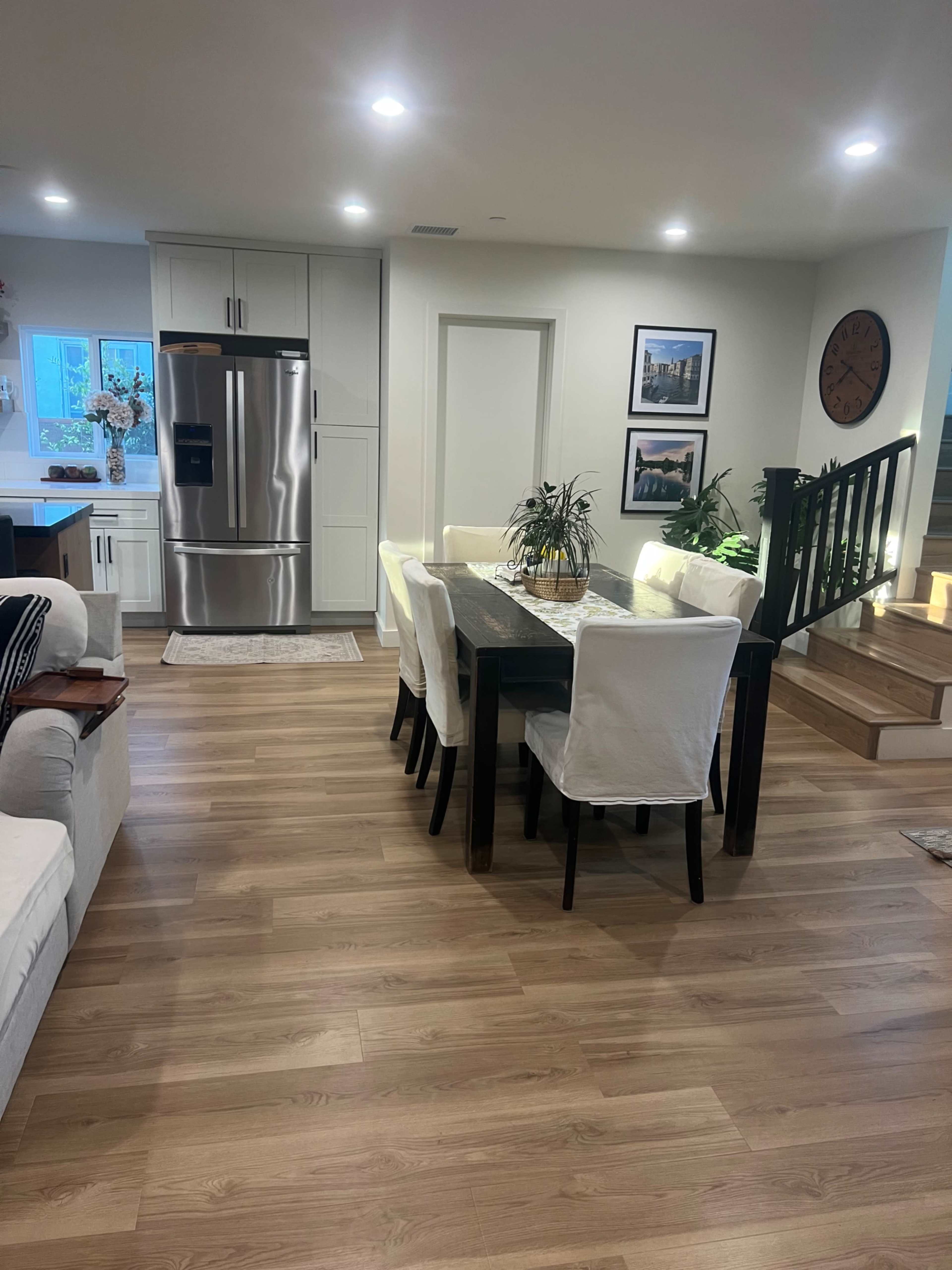 The image shows a modern dining area with a wooden table and white chairs, adjacent to a kitchen with stainless steel appliances and a staircase in the background.