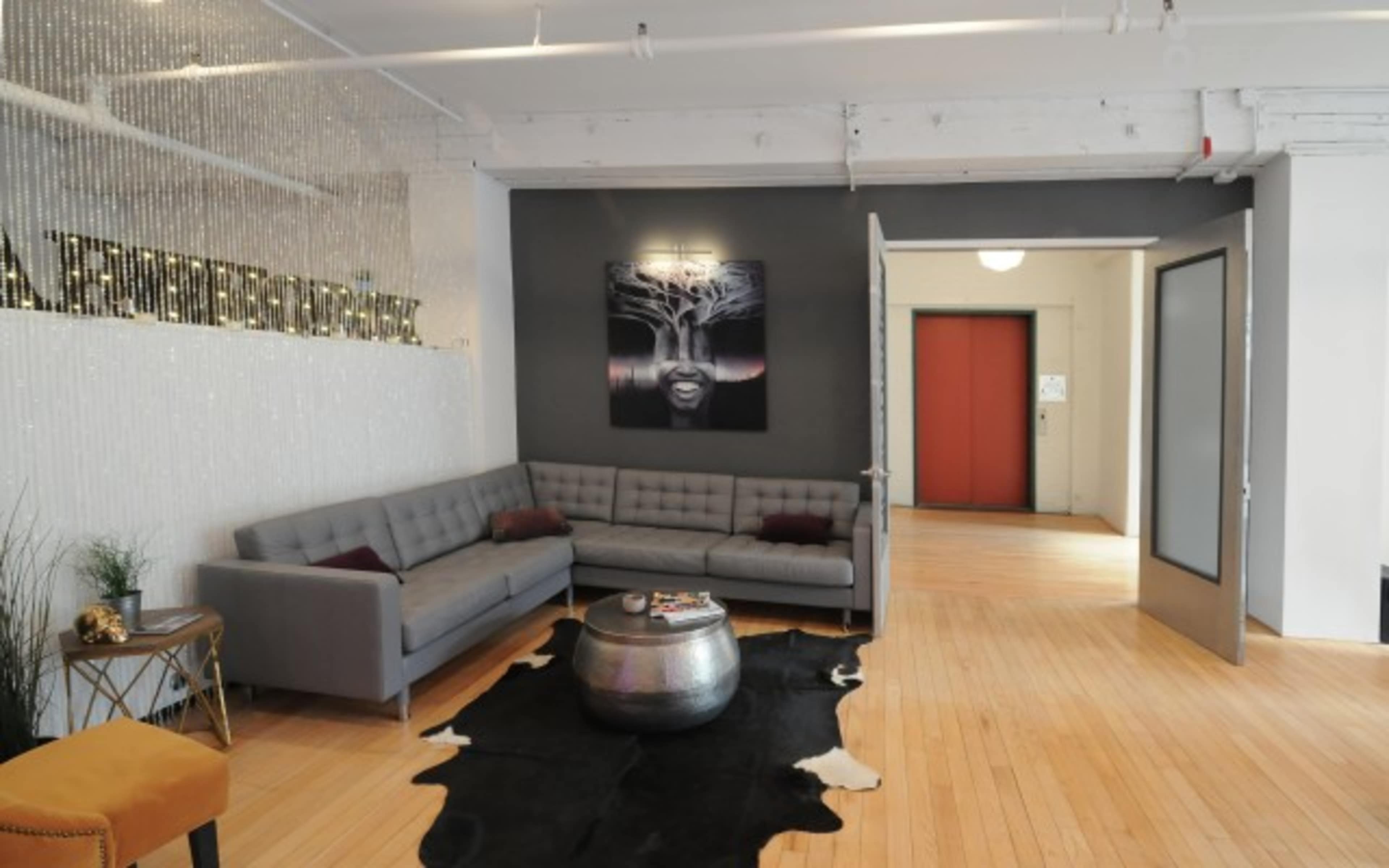 The image shows a modern lounge area with a sectional gray sofa, a round metallic coffee table, a black and white artwork on the wall, and an entrance leading to an elevator in the background.