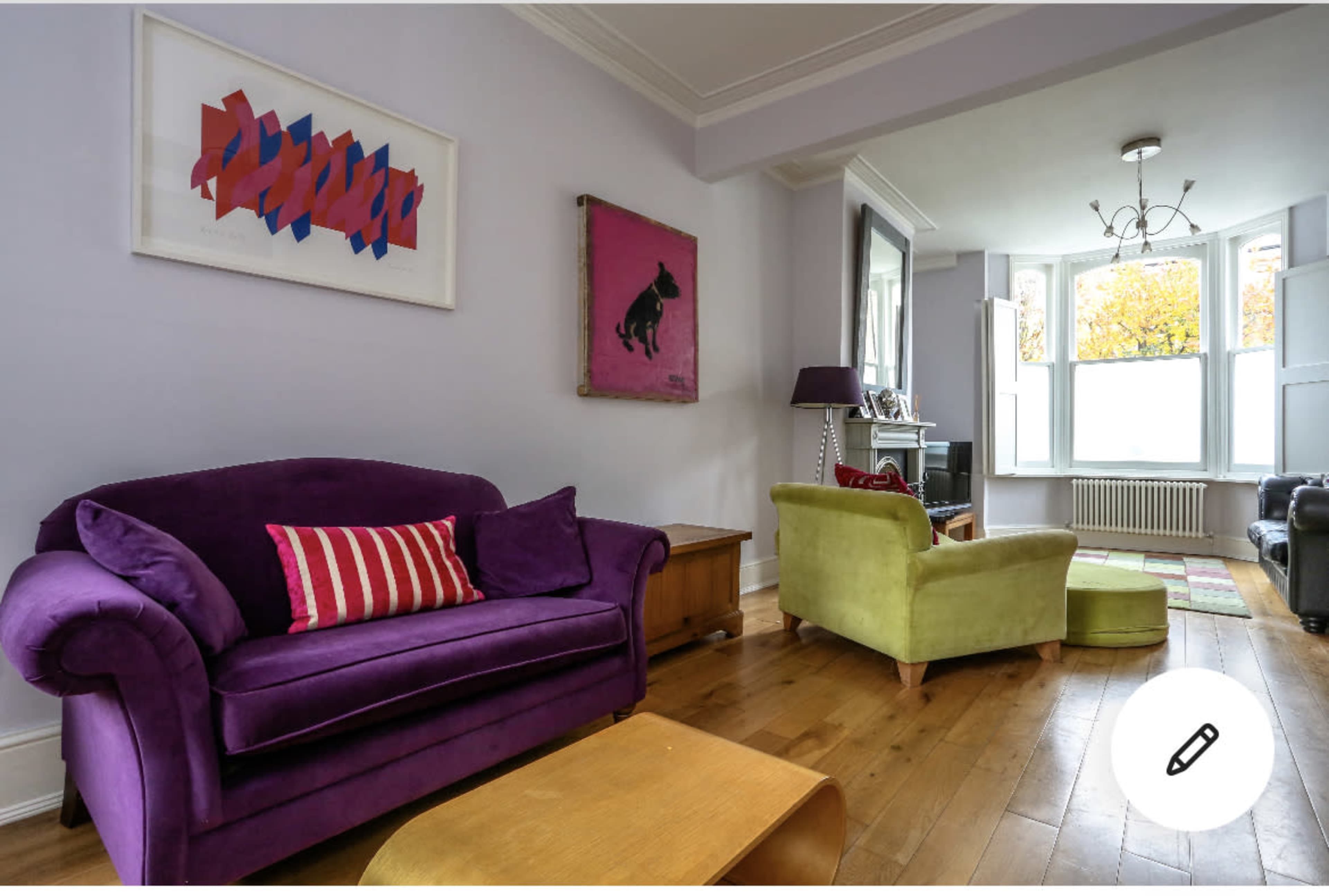 A living room with a purple sofa, a green armchair, and a wooden coffee table, along with artwork on the walls and large windows allowing natural light.