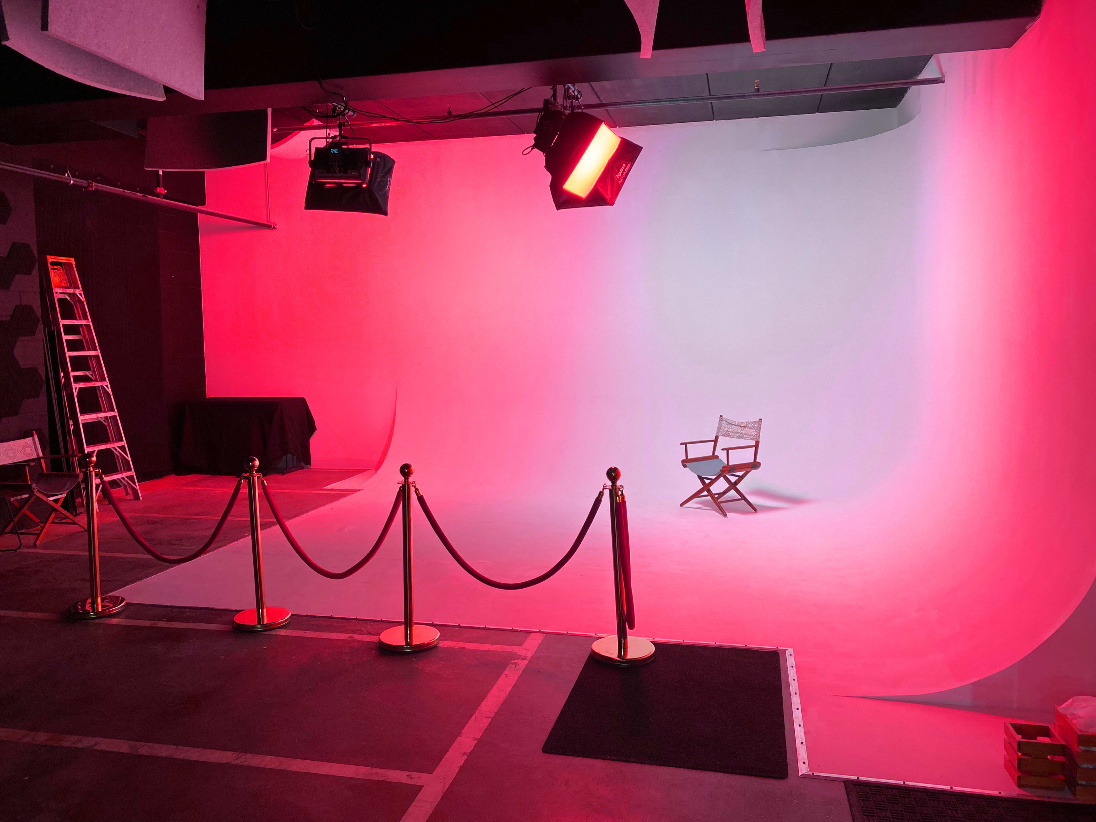 A photography studio features a pink and white backdrop, a director's chair, and a red spotlight, with a ladder and black table visible in the background.