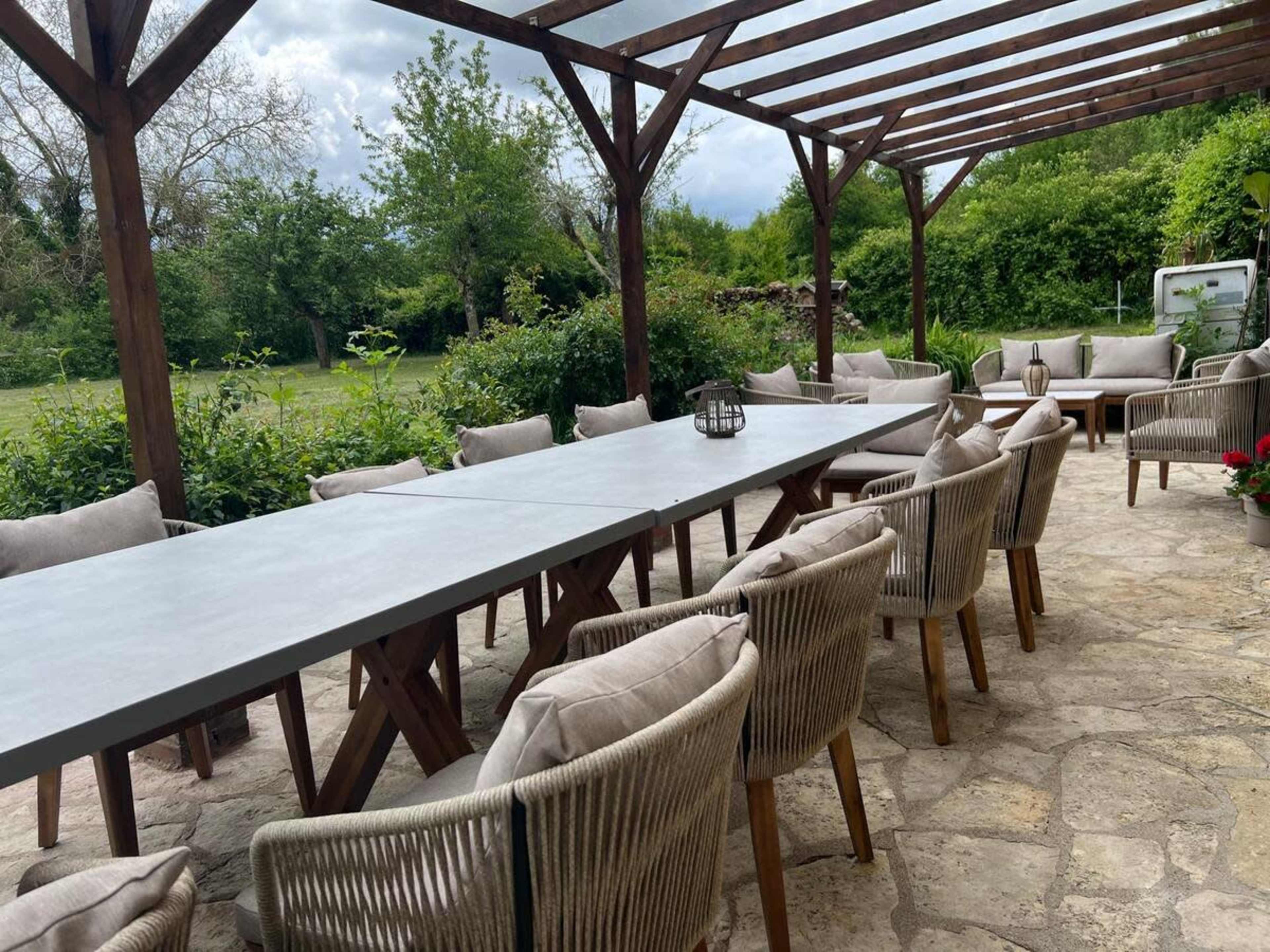 A long dining table with light-colored chairs is set under a wooden pergola in a garden area.