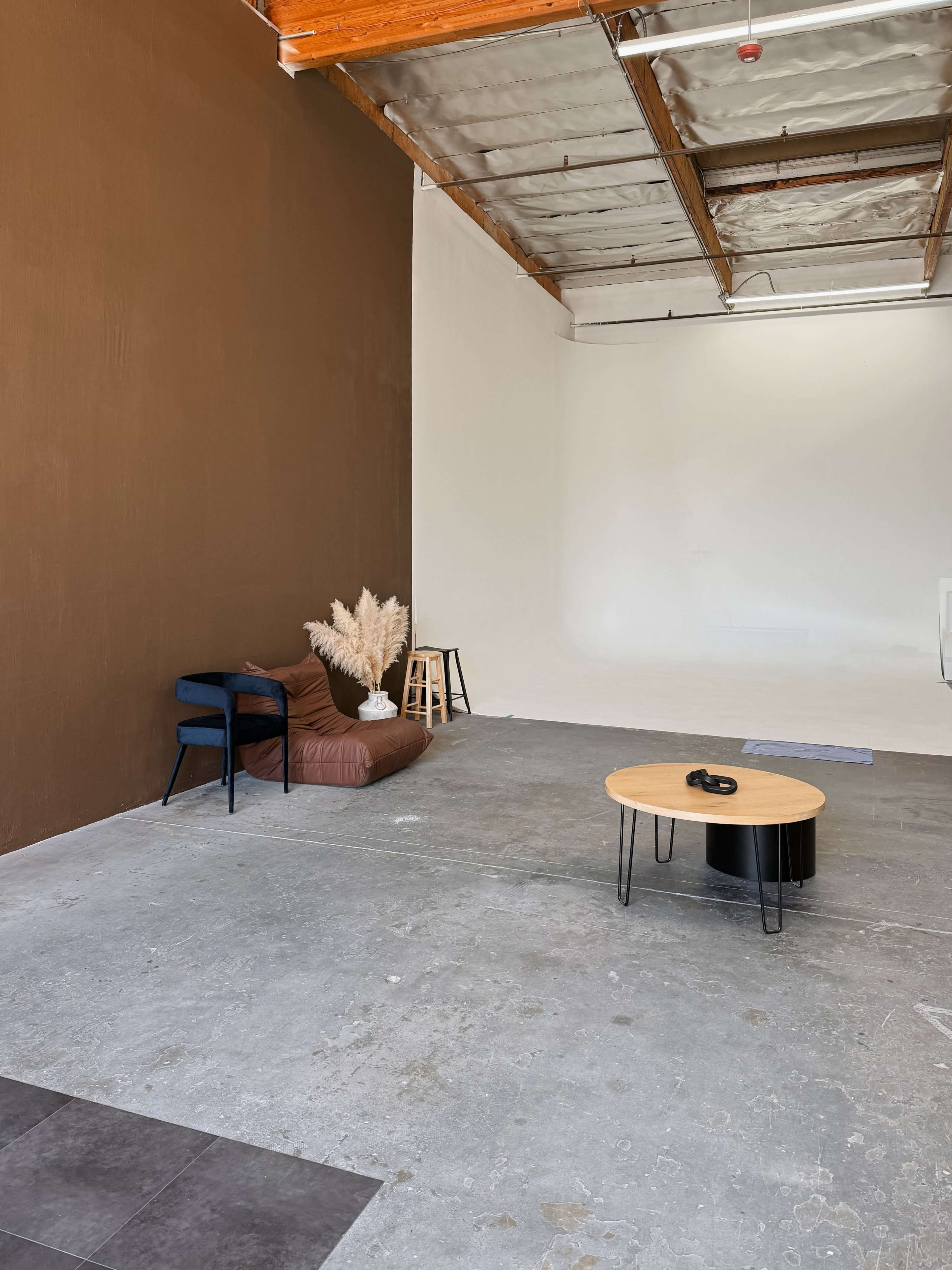 The image shows a minimalist studio space with a brown and white wall, a bean bag, a blue chair, a small black coffee table, and a stool.