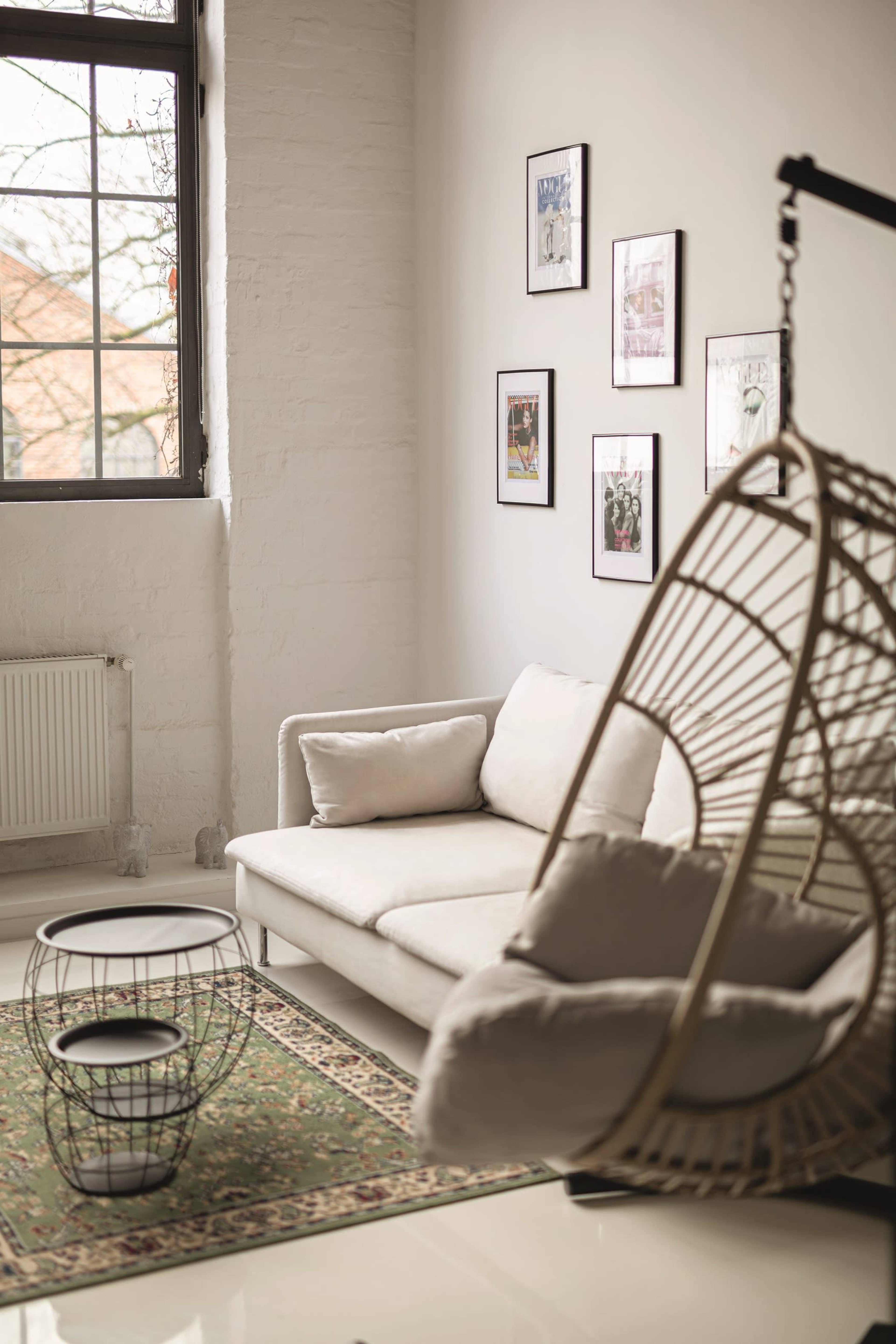 The image shows a modern living room with a white couch, a hanging chair, a small table, and framed pictures on the wall.