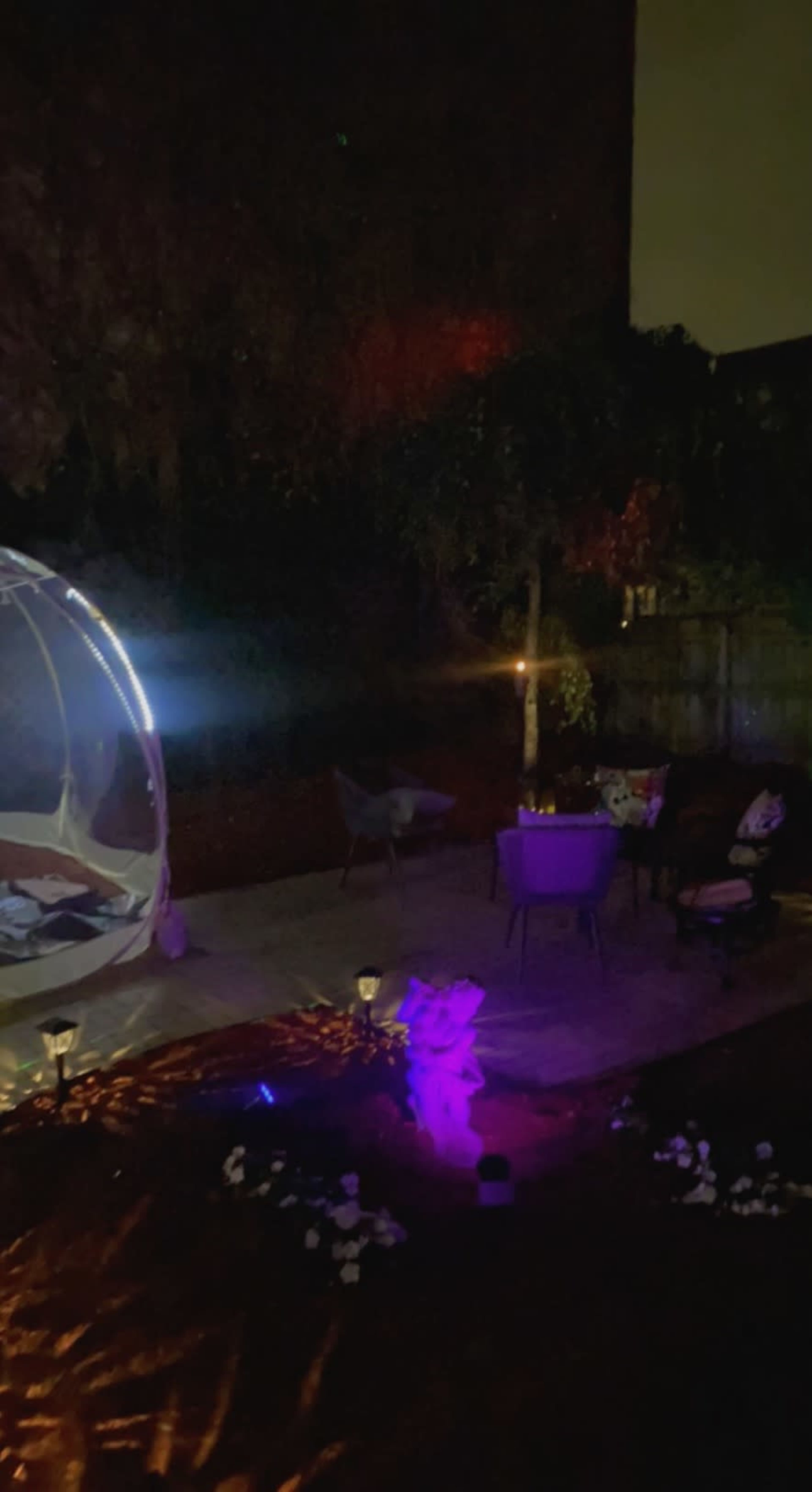 A dimly lit backyard features a hanging bed, a seating area with chairs, and colorful lights illuminating the space.