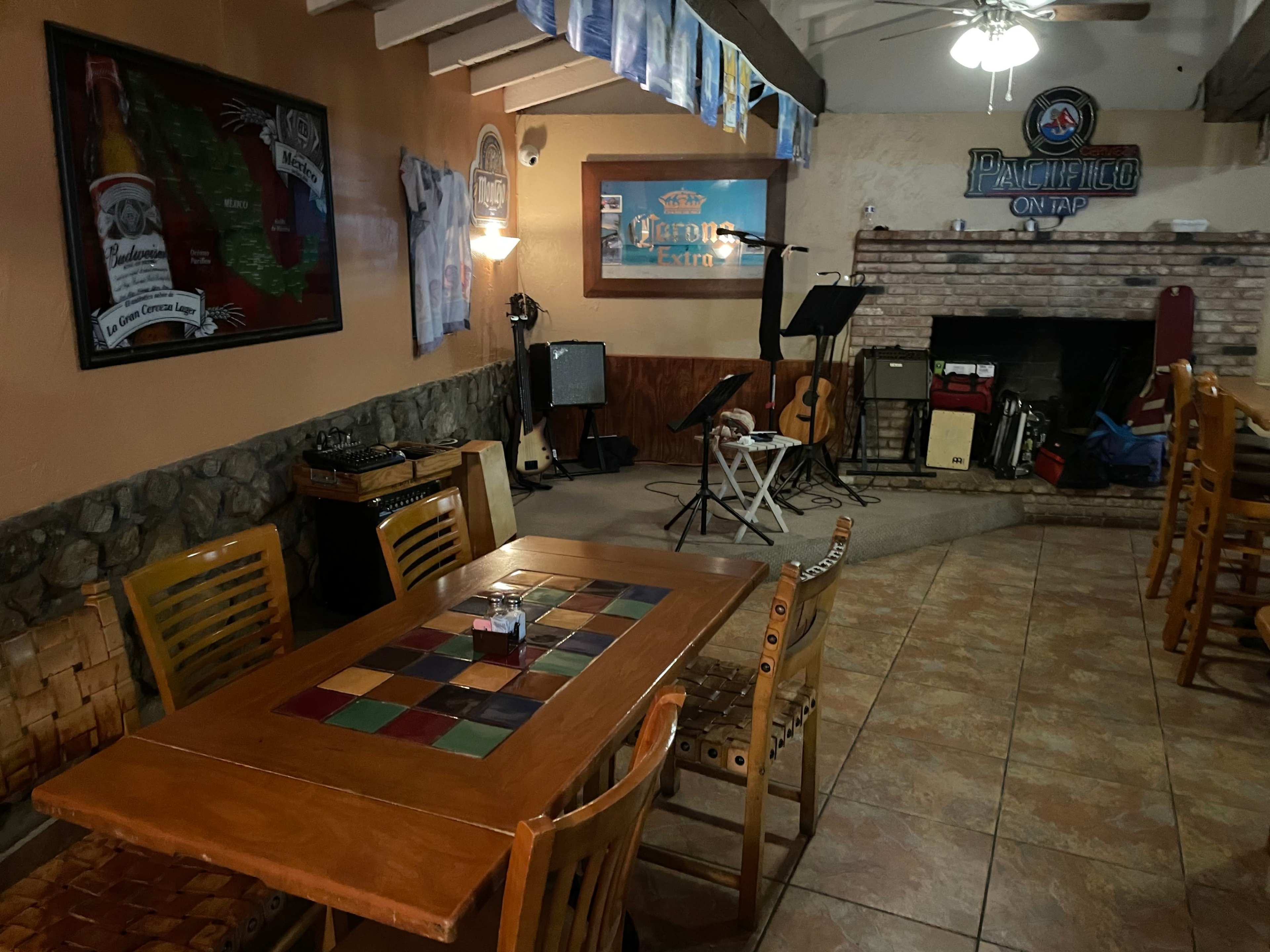 An empty restaurant with a colorful table, a small stage set up for performances, and musical instruments at the back near a stone fireplace.