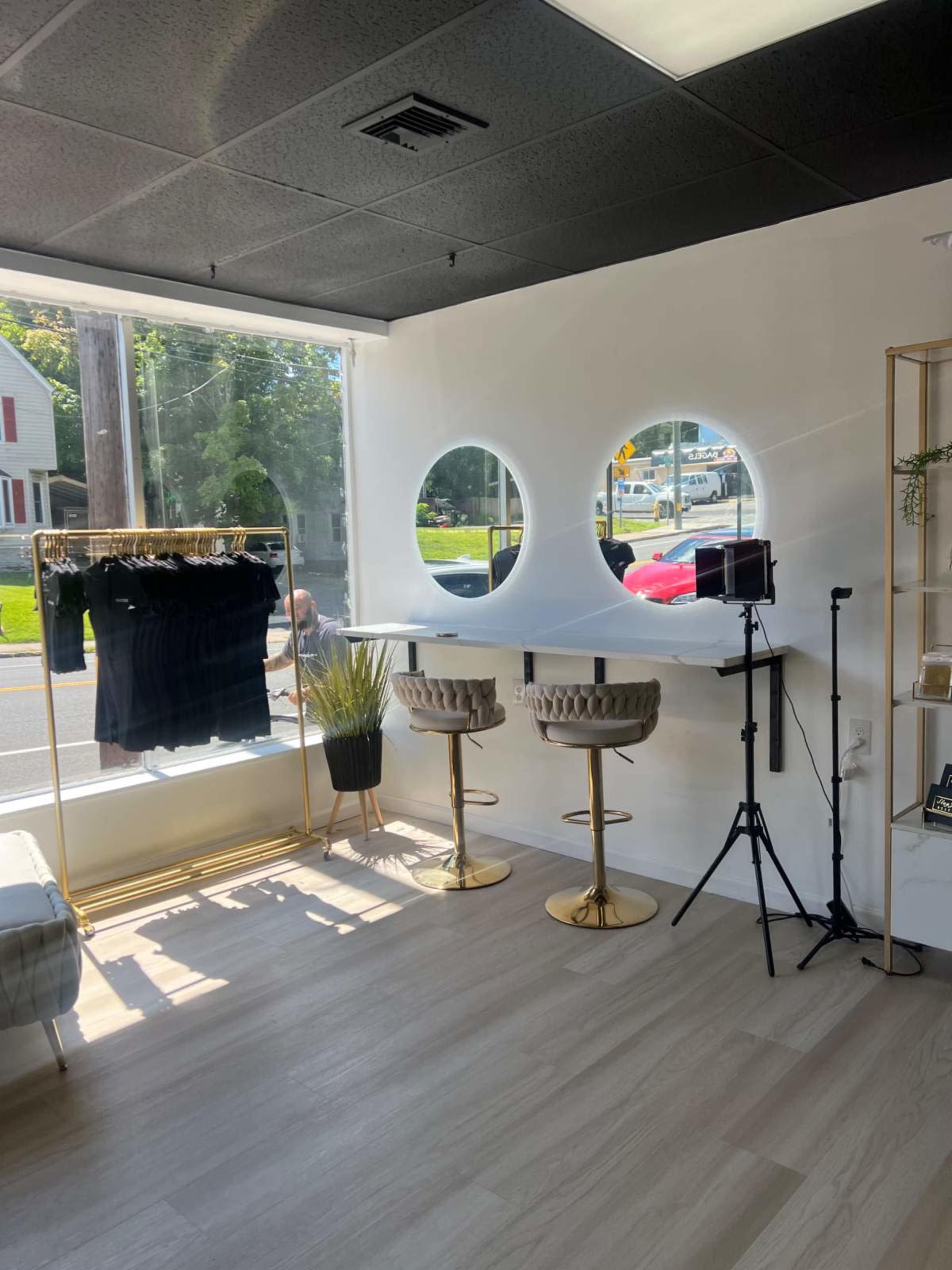 A modern retail interior featuring two circular windows, gold-accented bar stools, a clothing rack with black garments, and a small backdrop setup for photography.