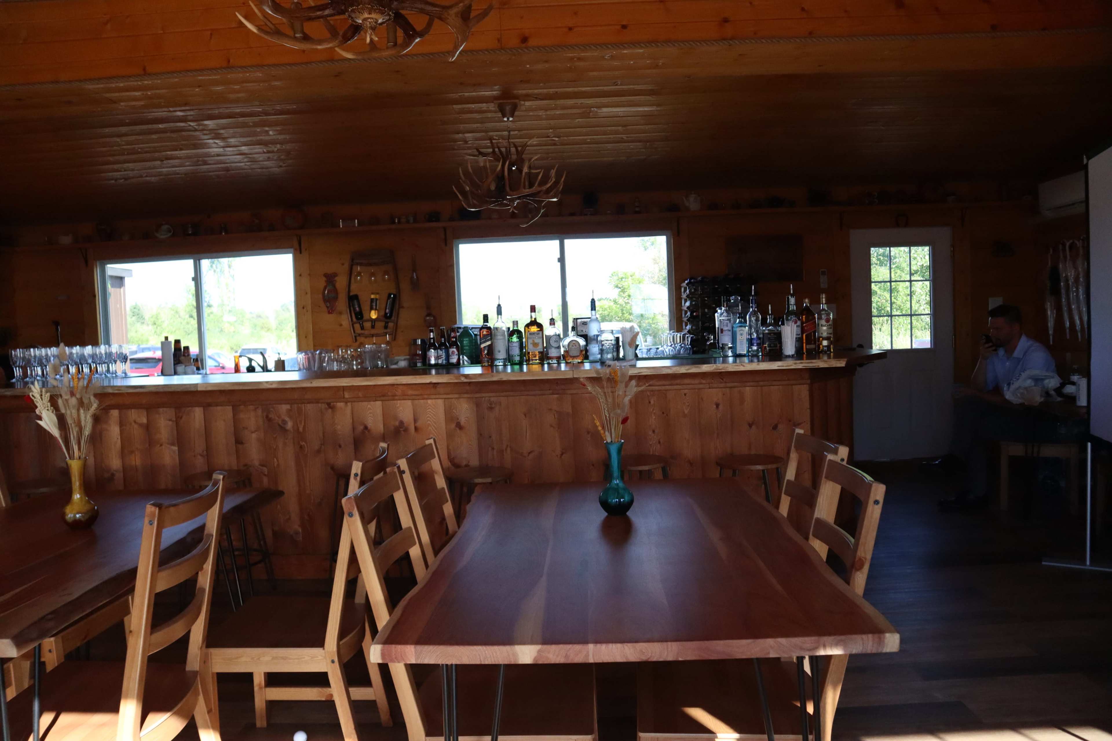 The interior of a wooden bar area with a long counter, shelves of various liquor bottles, and several dining tables arranged around the space.