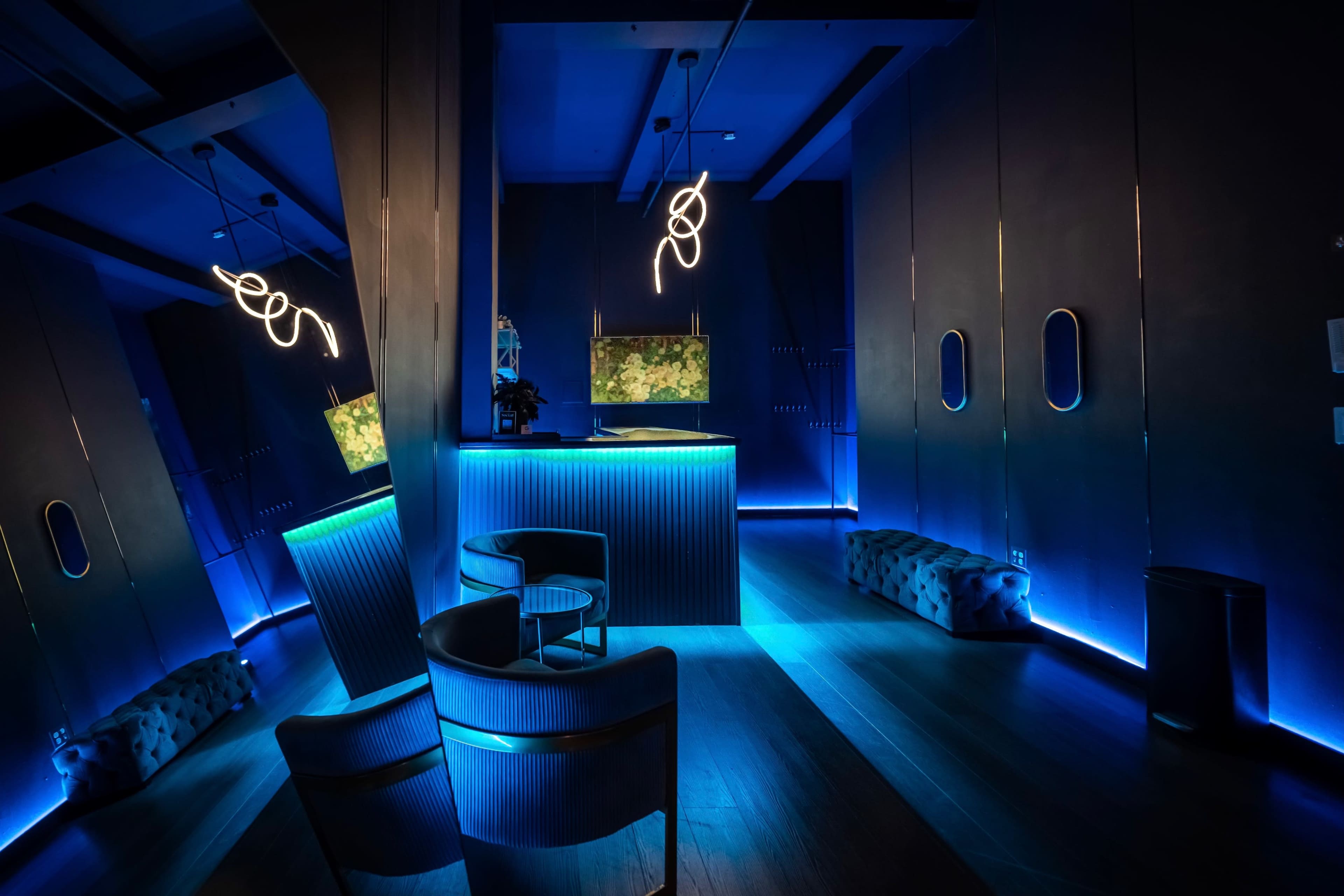 The image depicts a modern interior space with dim lighting, featuring sleek furniture, a glowing bar, and decorative lighting elements along the walls.