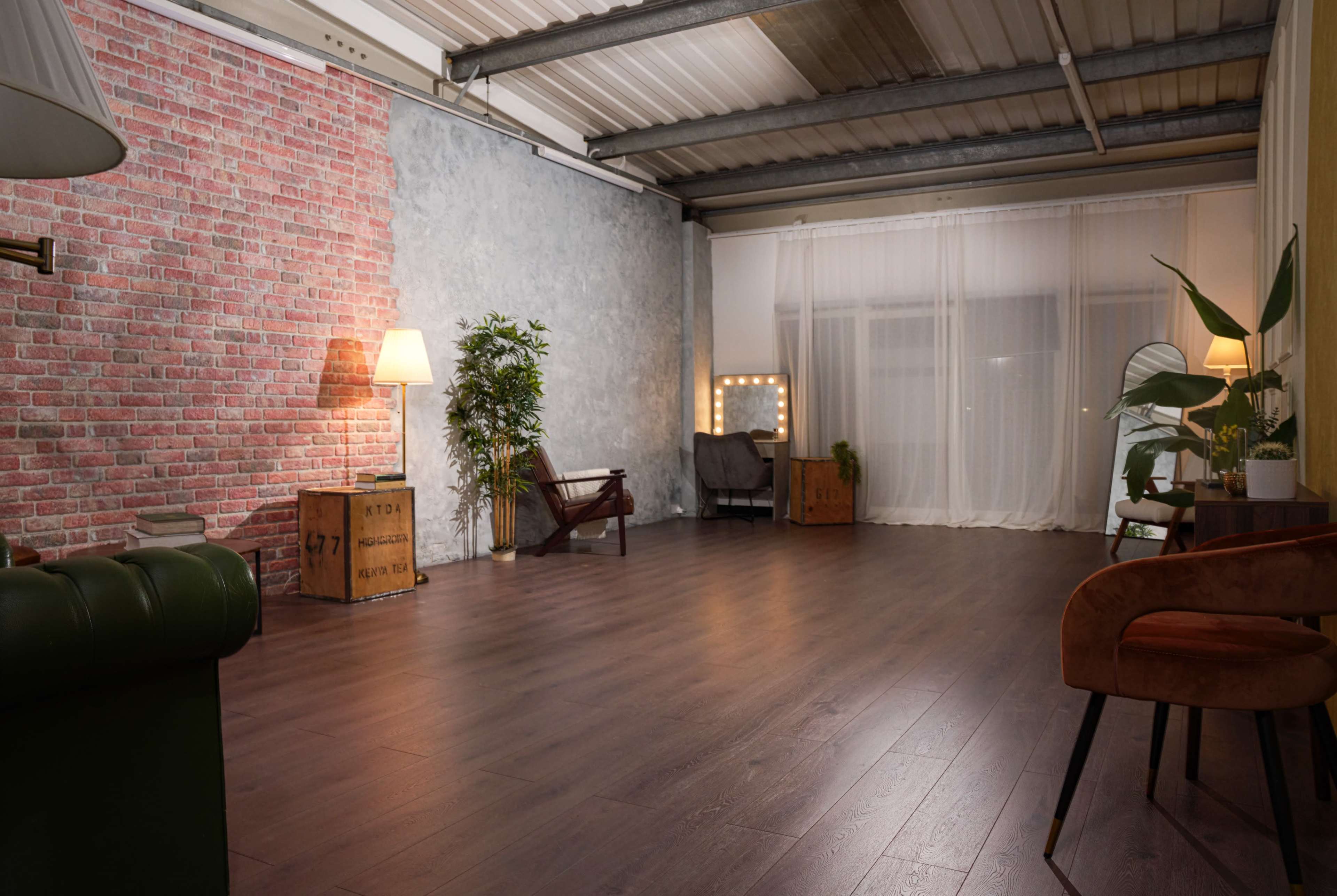 The image shows a spacious interior with a mix of exposed brick and concrete walls, wooden flooring, various plants, and several seating areas, including chairs and lamps.
