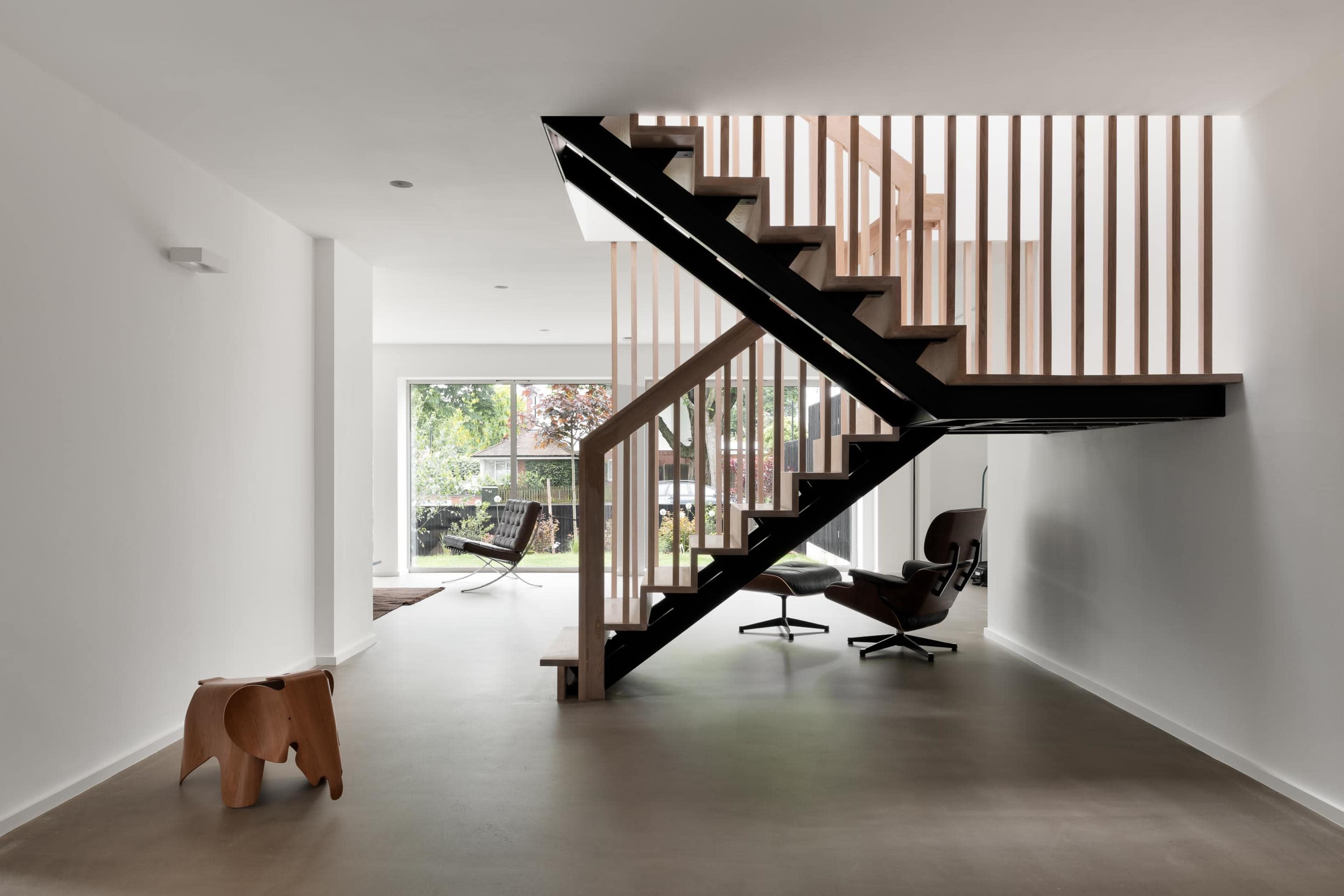 A modern interior space featuring a wooden staircase with a railing, a minimalist elephant-shaped stool, and a view of outdoor seating through a large glass door.