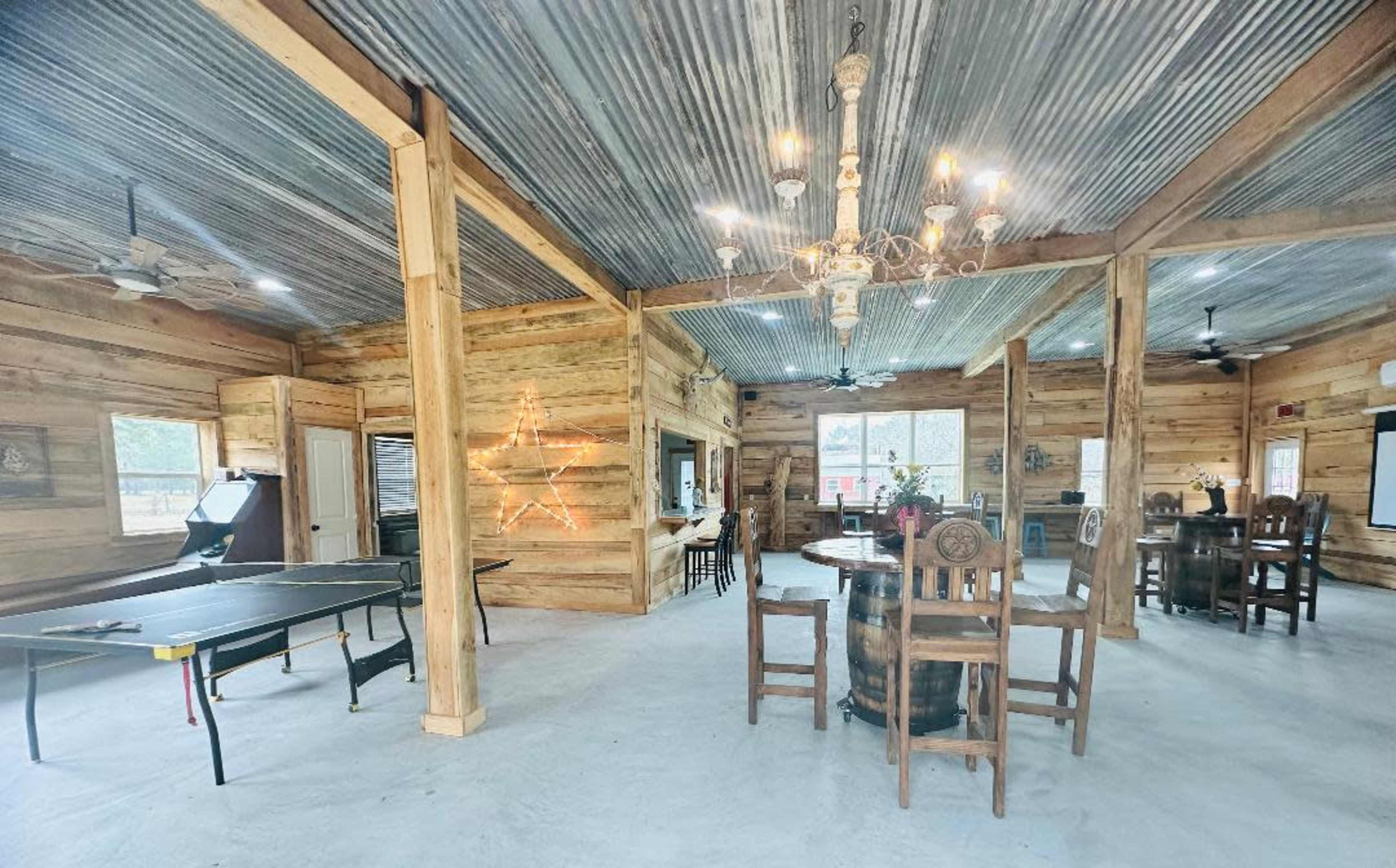 A spacious interior with wooden walls, a ping pong table, and a dining area featuring a large wooden table and chairs, illuminated by a chandelier.