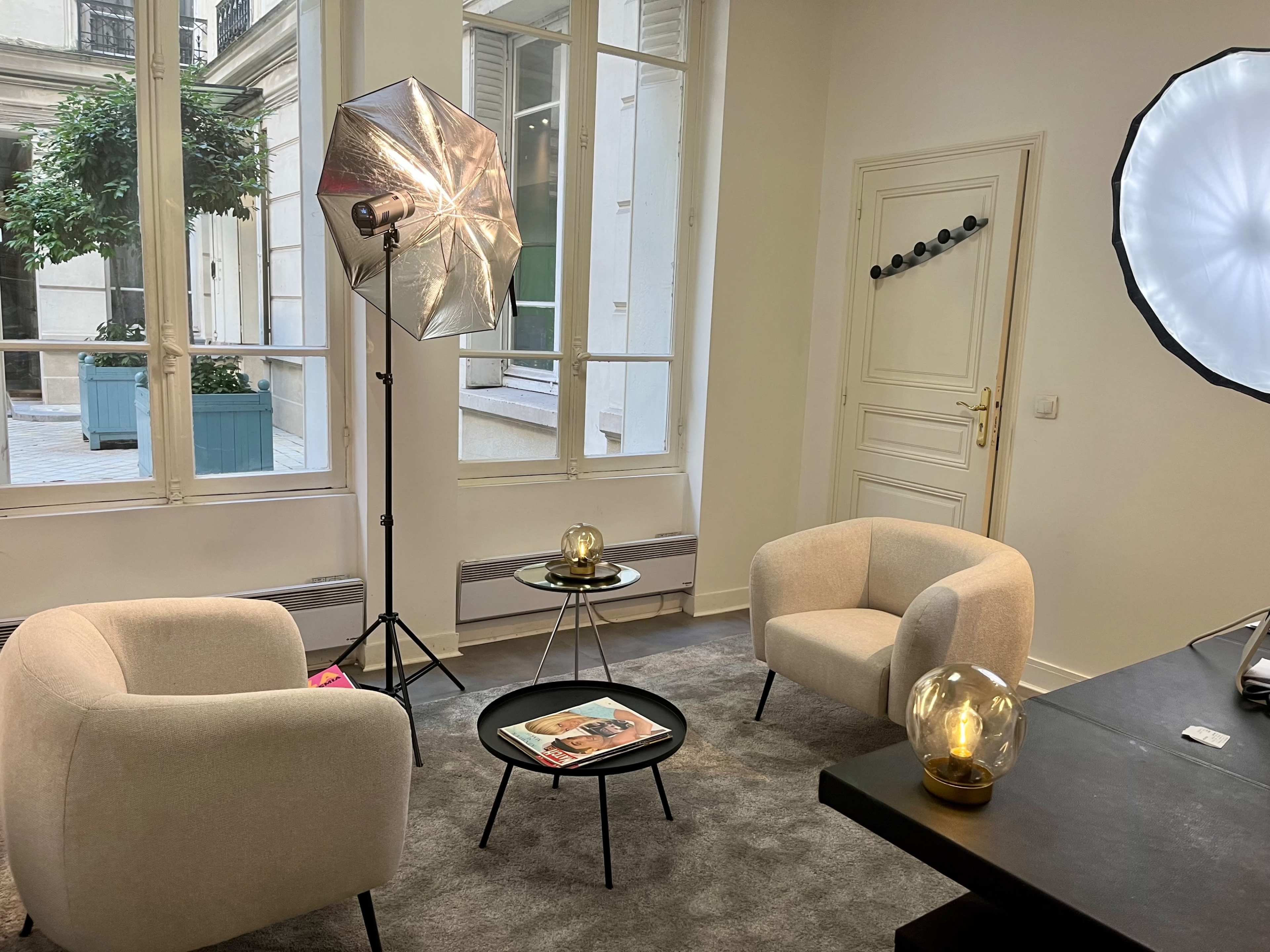 A well-lit interior space featuring two beige armchairs, a small black coffee table with a magazine, and photography equipment in front of large windows.