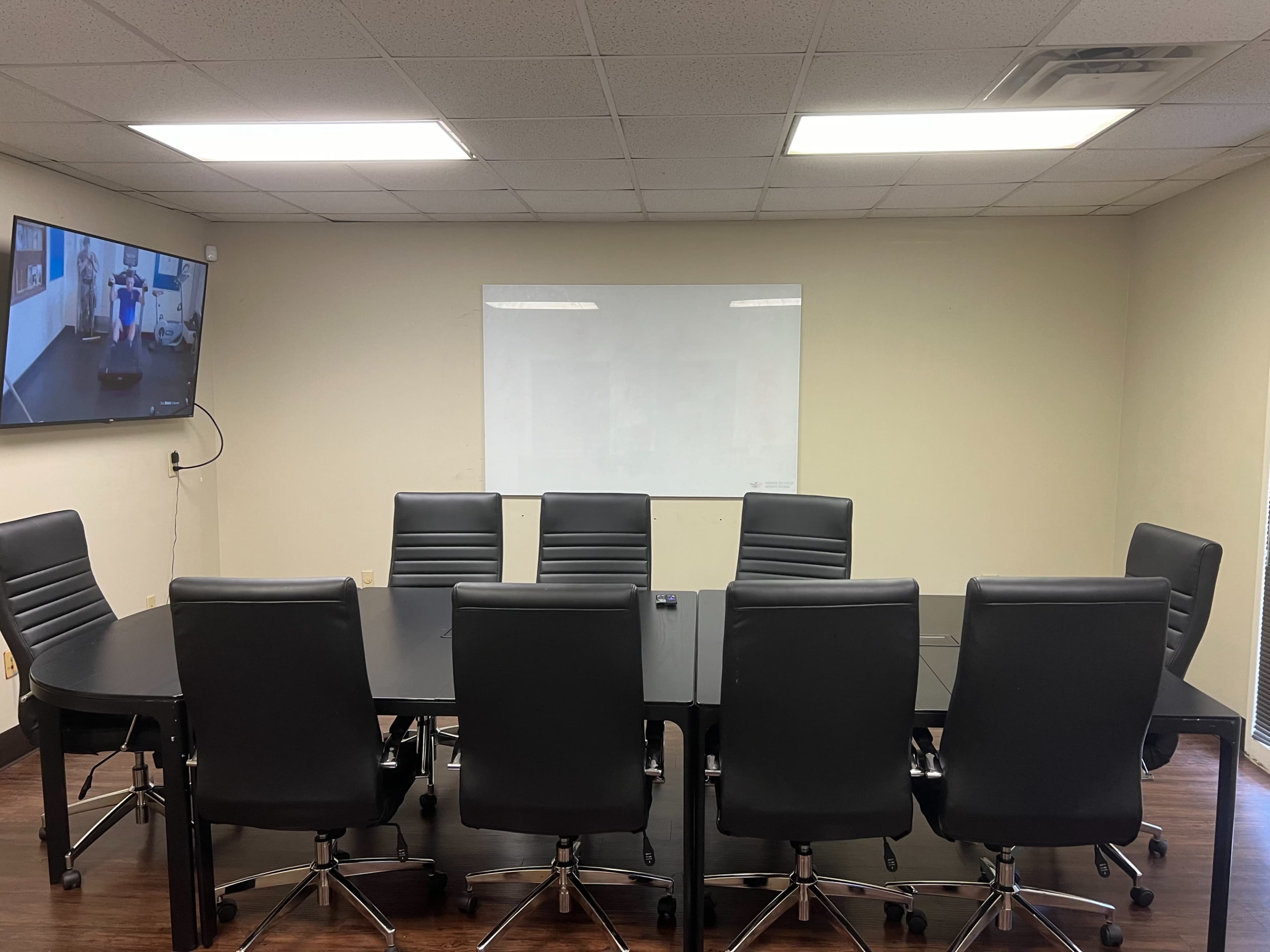 A conference room with a large black table surrounded by eight black leather chairs and a wall-mounted screen displaying a video.