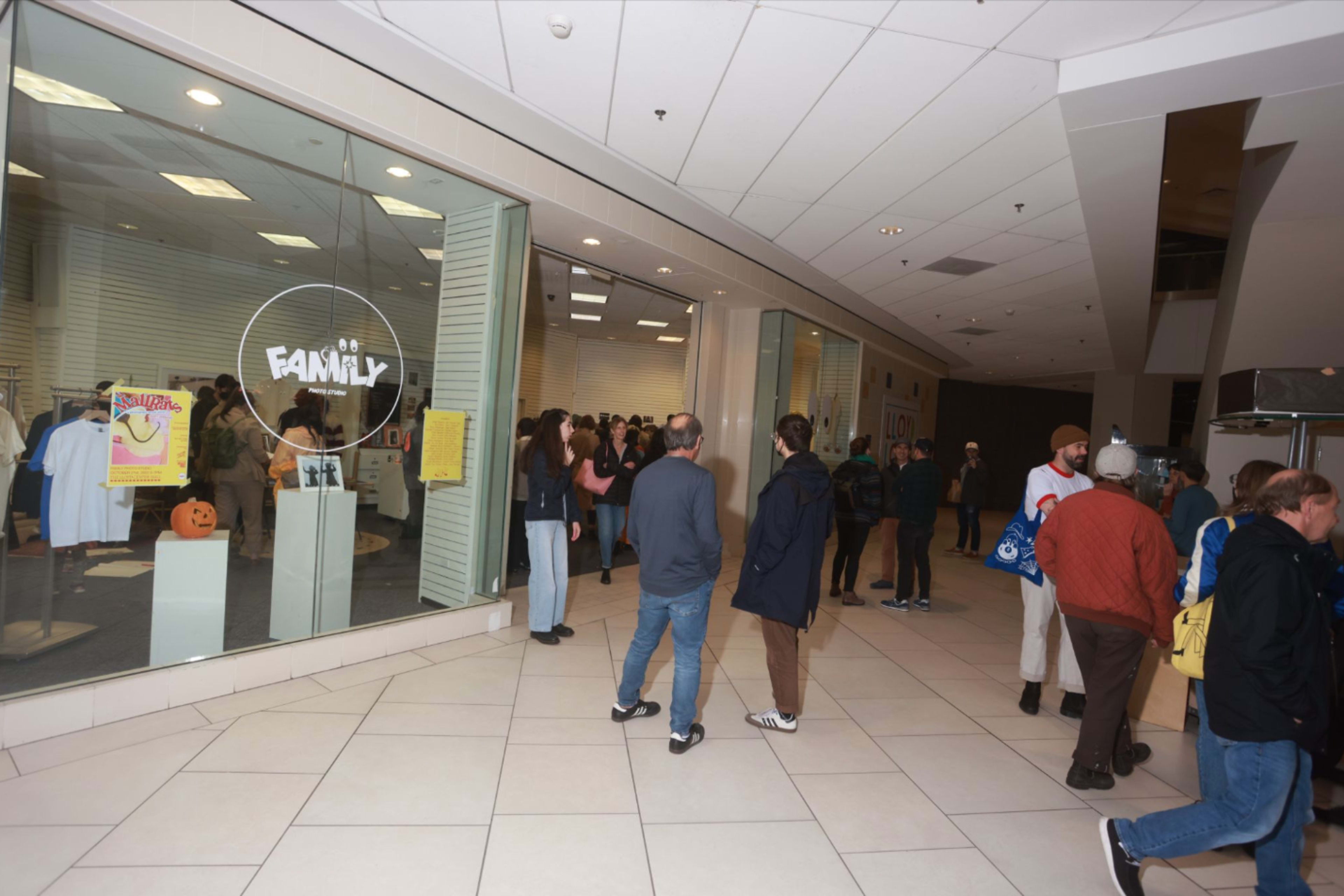 A group of people gathers in a shopping mall near a store called "FAMILY," with windows displaying clothing and decorations.