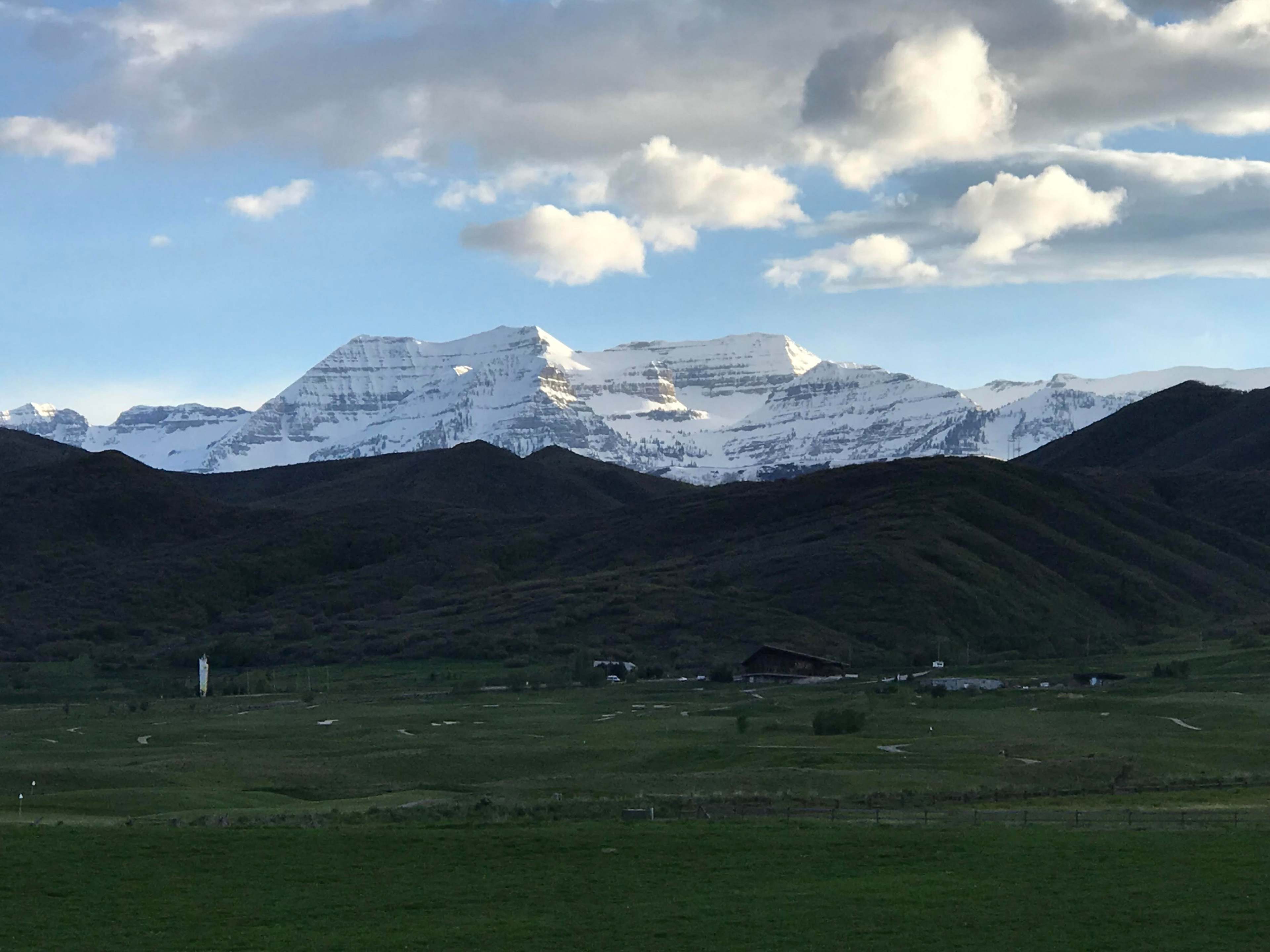 A mountainous landscape with snow-capped peaks rising above green hills and a valley, under a partly cloudy sky.