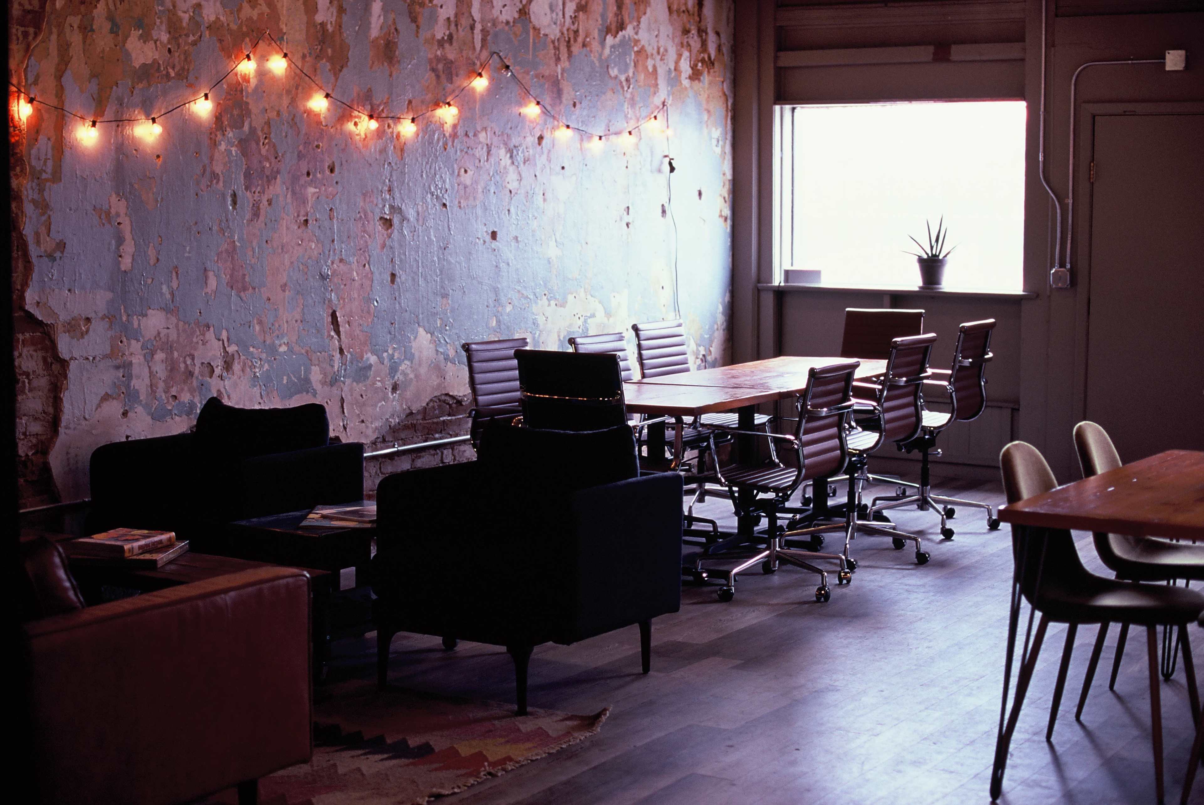 A workspace with several modern chairs and tables arranged against a wall with peeling blue paint, illuminated by string lights.