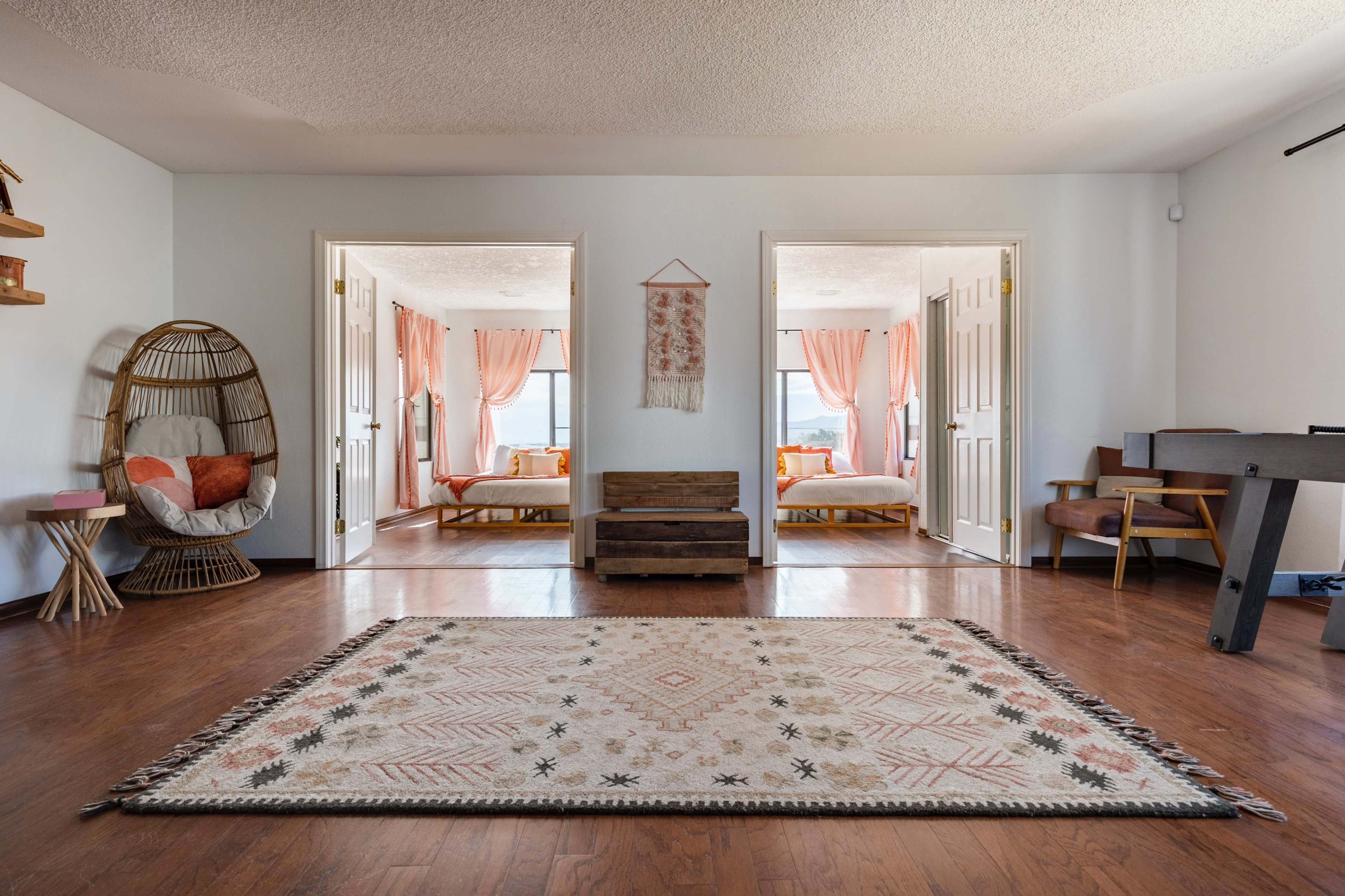 A spacious room with wooden flooring features two doorways leading to brightly lit bedrooms, a patterned area rug in the center, and a mix of decorative furniture on one side.