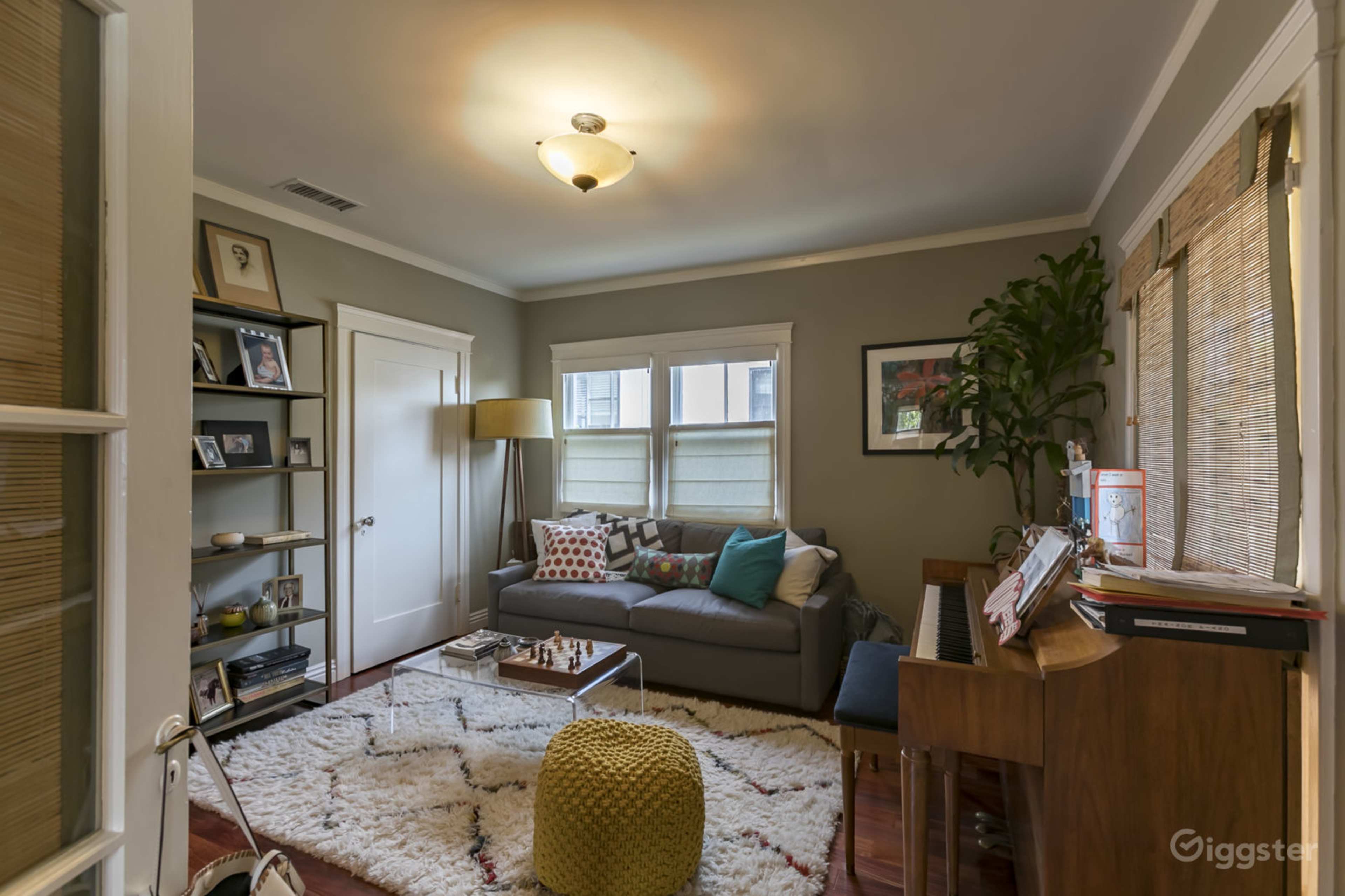 The image shows a cozy living room with a gray sofa, a bookshelf, a piano, and a large white rug on the wooden floor.