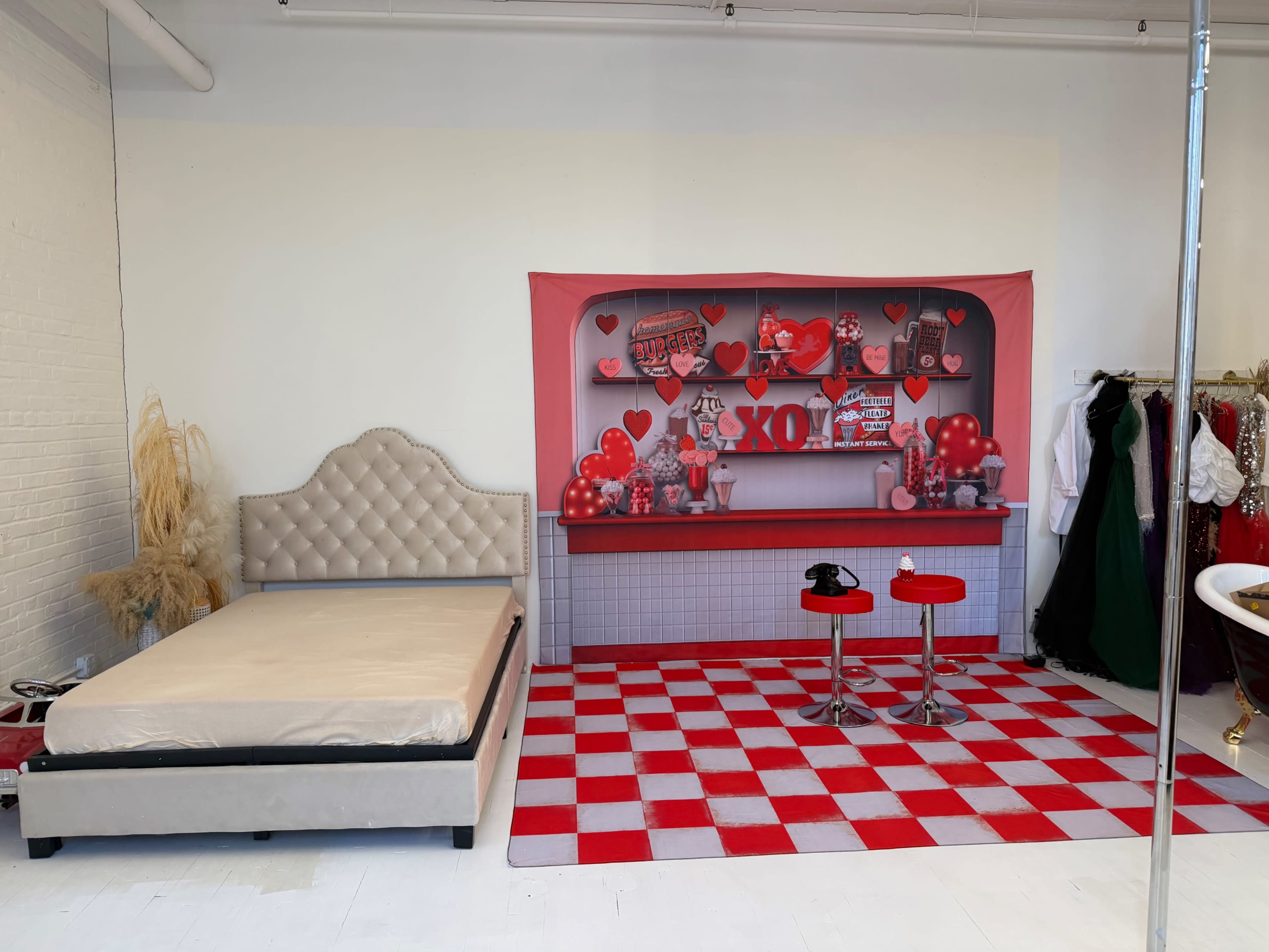 The image shows a room with a beige bed, a red and white checkered floor, and a backdrop featuring a shelf decorated with hearts and balloons.