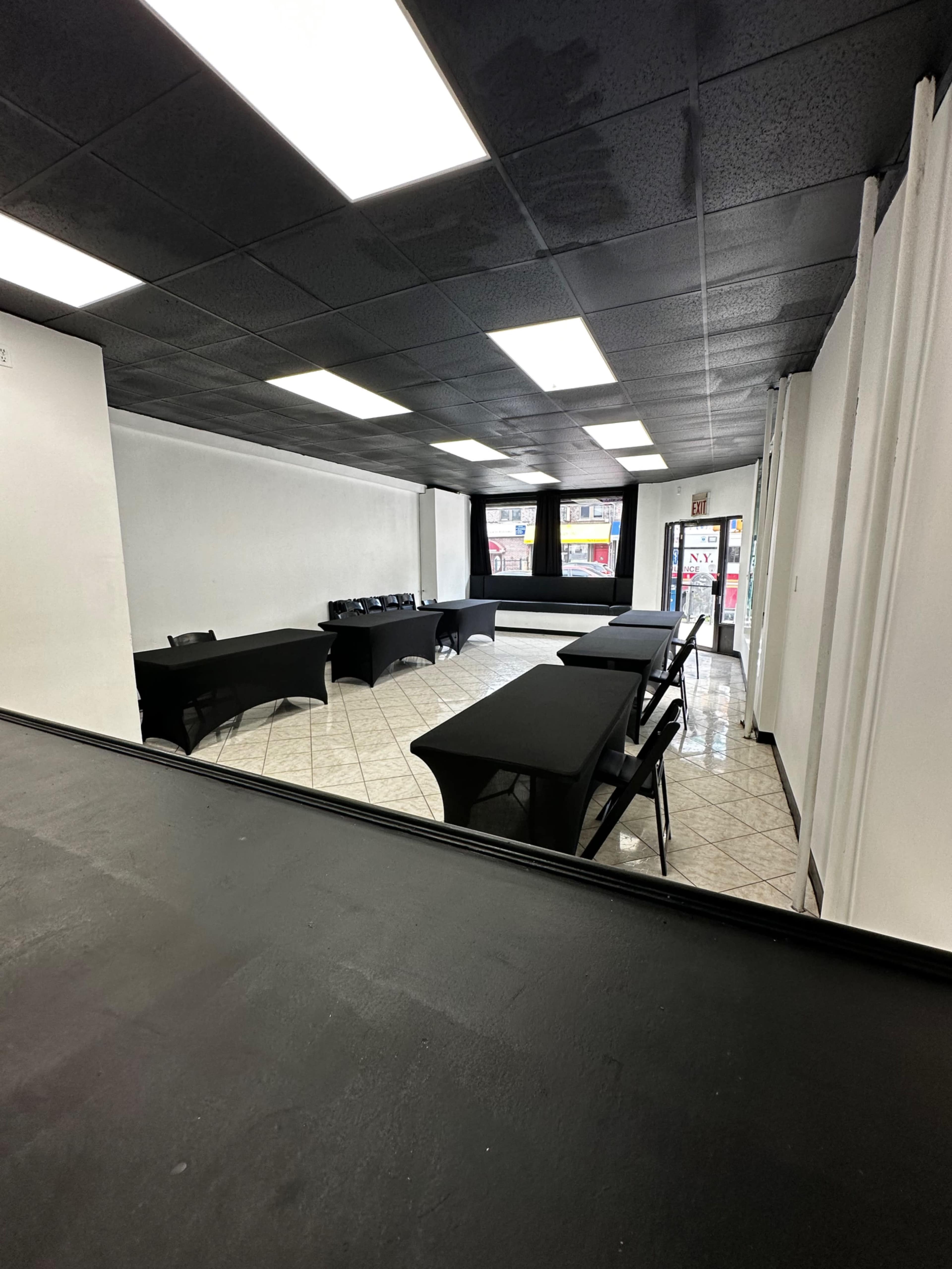 The image shows a sparsely furnished room with black tables arranged in a U-shape on a tiled floor, and a door leading outside at one end.