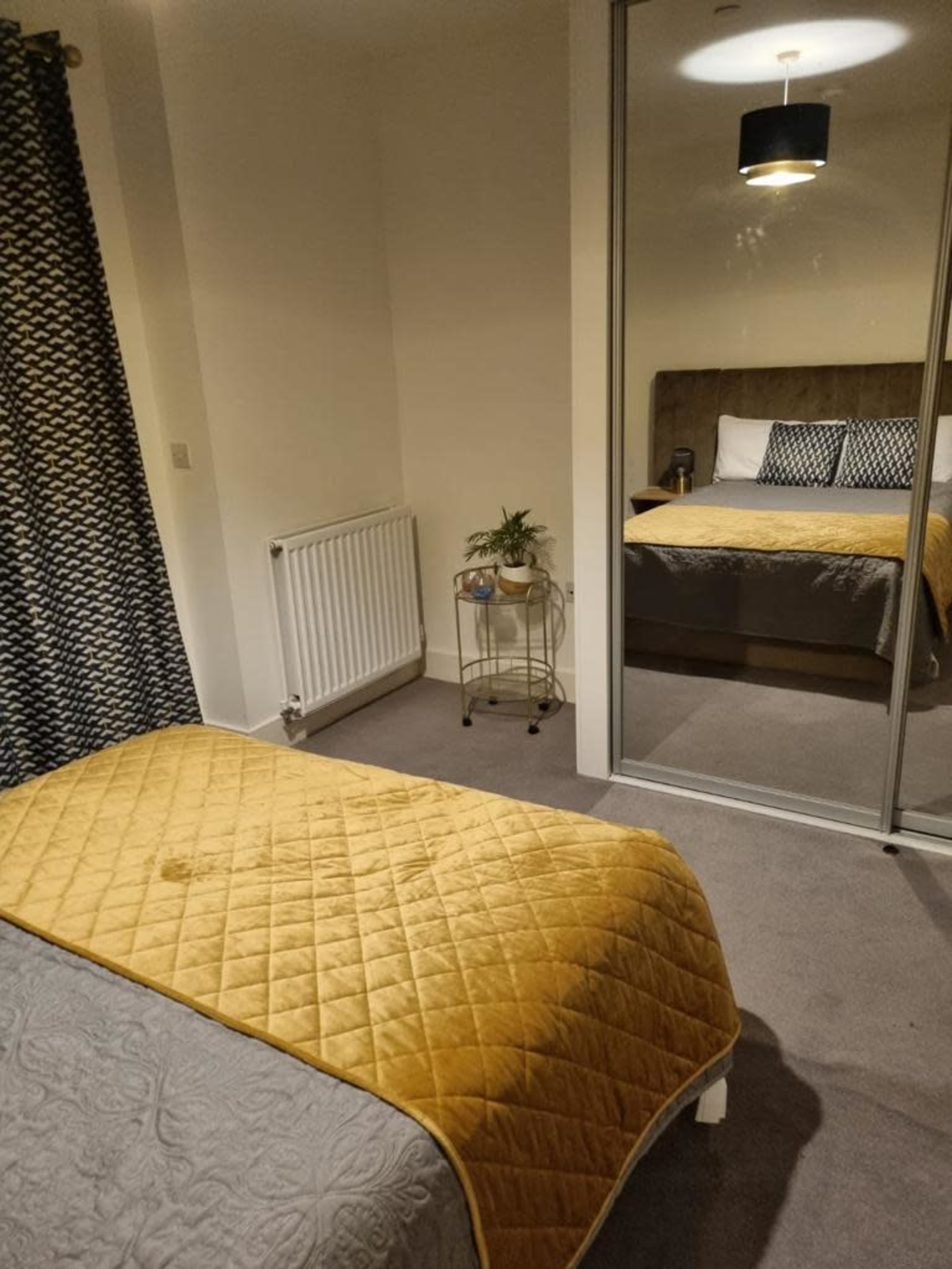 The image shows a bedroom with two beds, a mirror reflecting the main bed, a small plant on a bedside table, and a radiator on the wall.