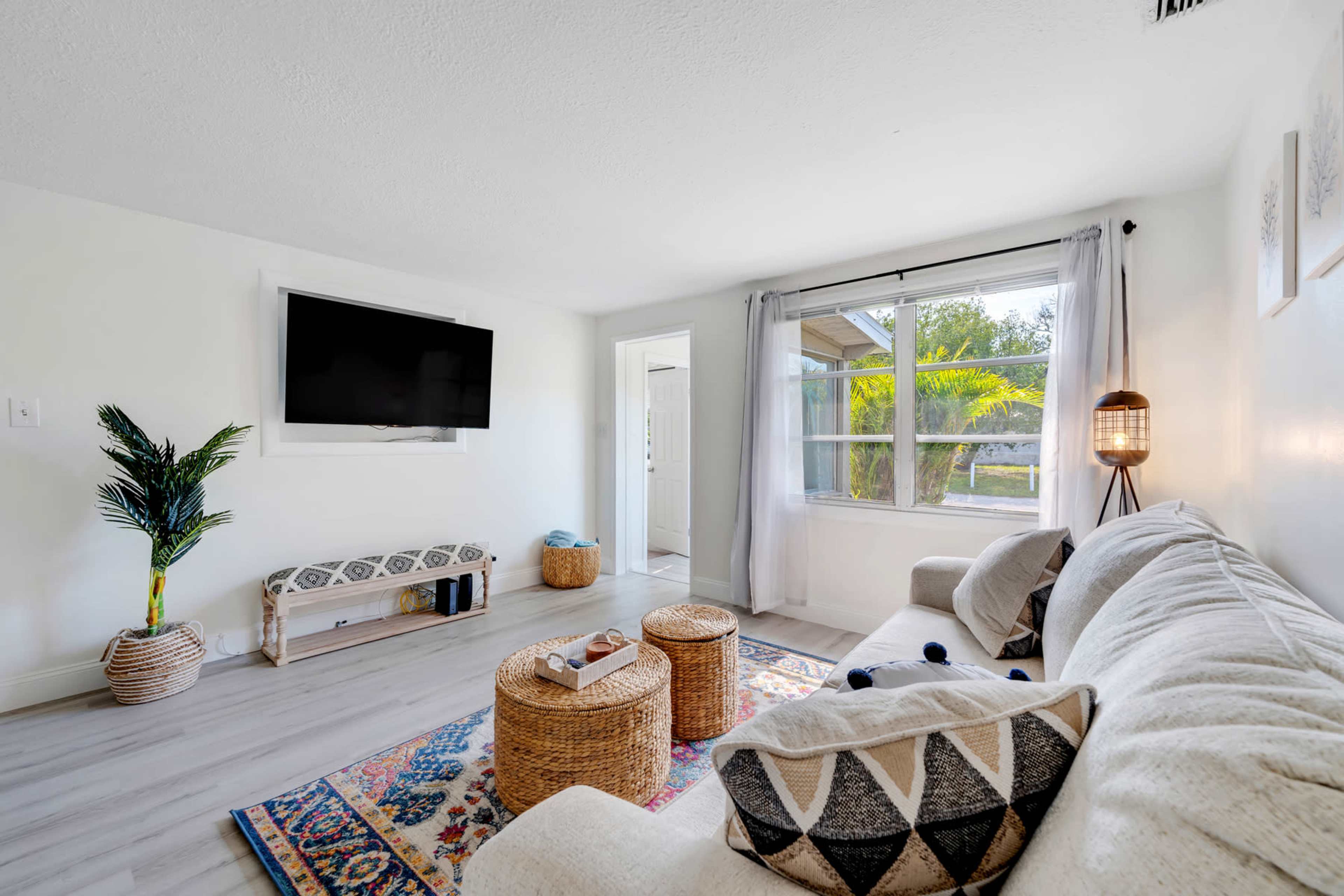 A bright living room with a large window, a comfortable sofa, a television mounted on the wall, and two round wicker tables.