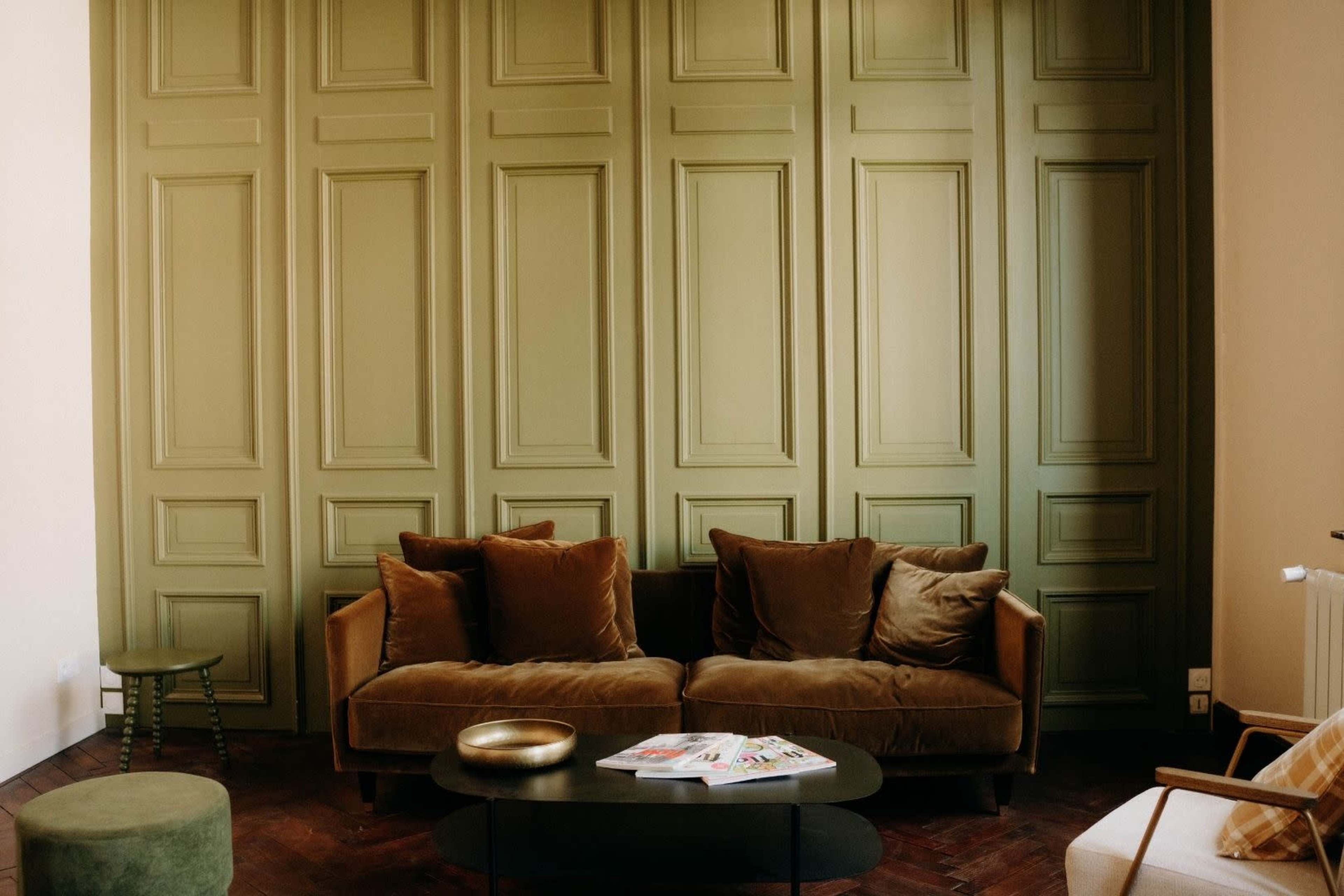 The image shows a cozy living room featuring a green paneled wall, a brown velvet sofa, a black coffee table, and a small green side table.