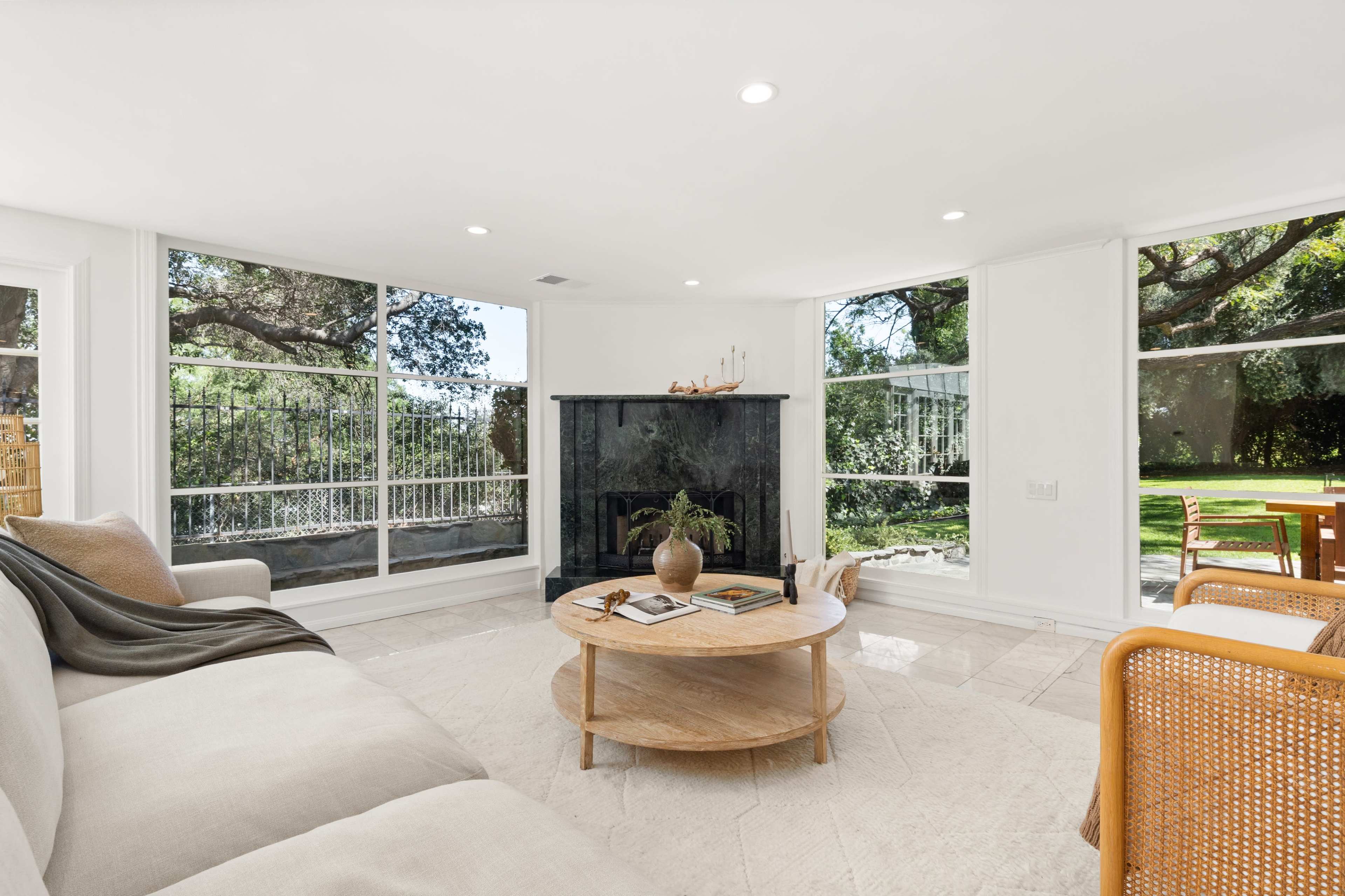 A modern living room features large windows overlooking a green yard, a black fireplace, a round wooden coffee table, and a comfortable sofa.