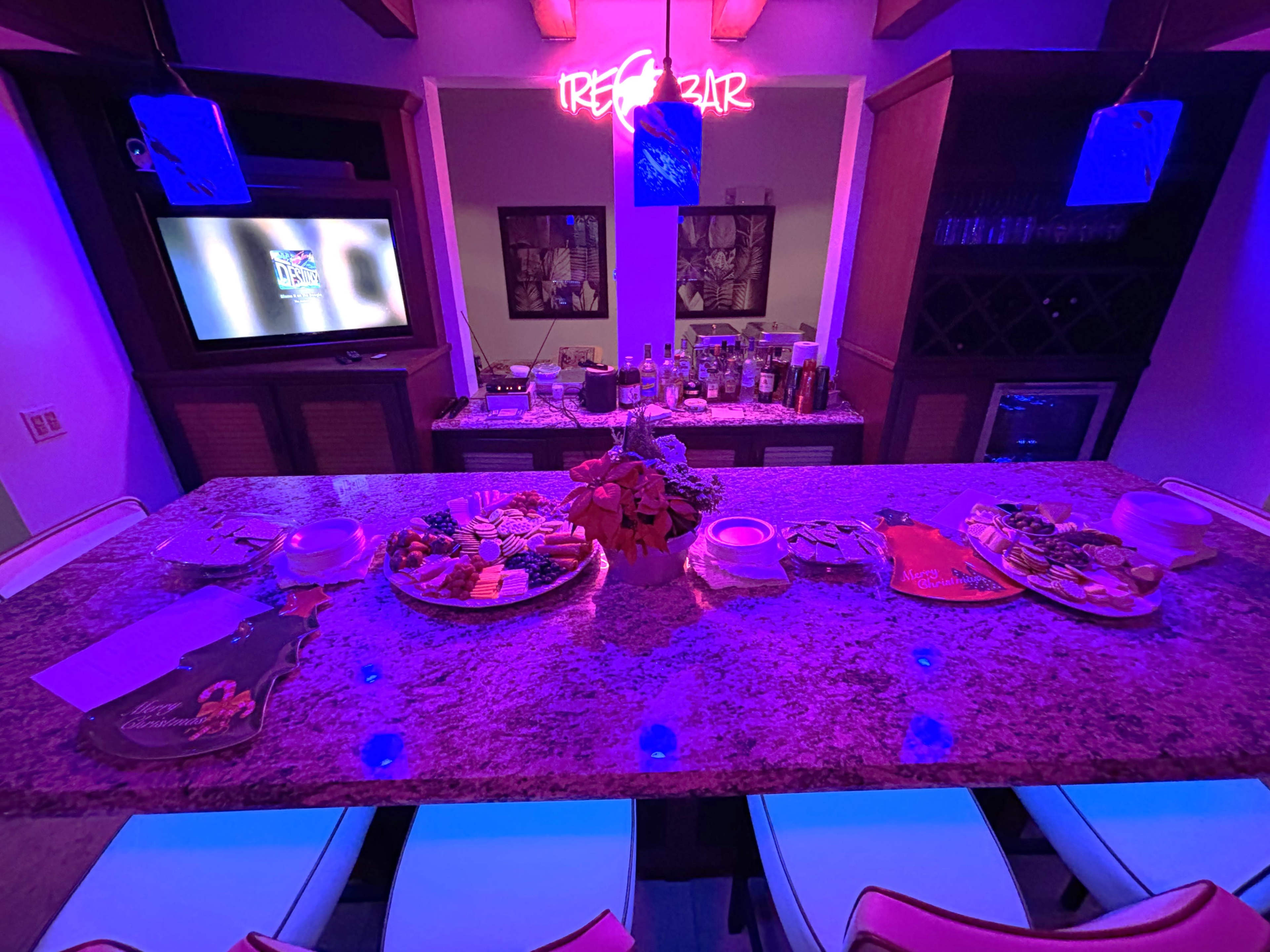 A well-lit bar area features a granite countertop displaying various appetizers and drinks, with a neon "TREZ BAR" sign in the background.