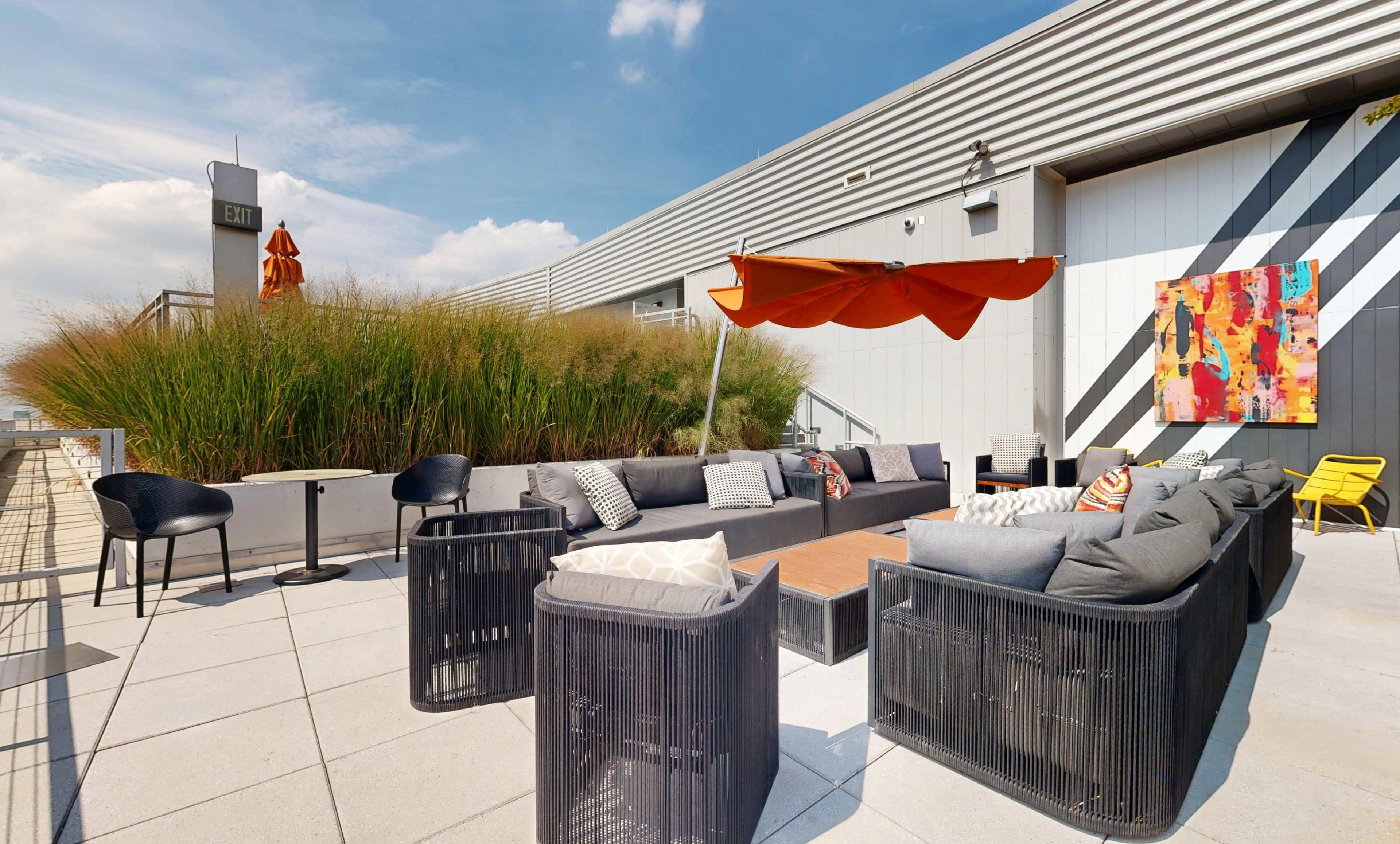 The image shows a rooftop patio featuring modern seating arrangements, a large orange umbrella, and an abstract painting on the wall.