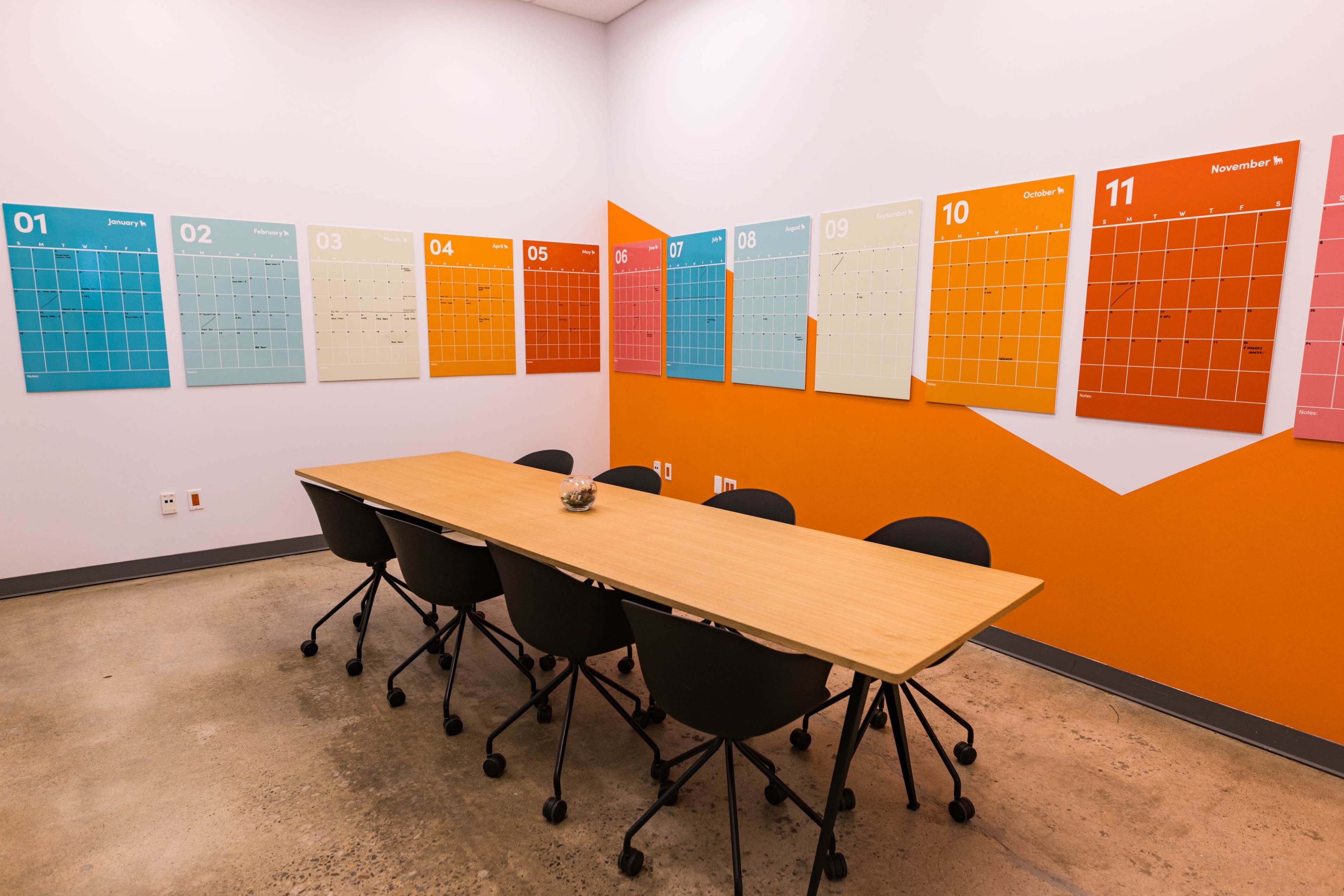 A conference room features a long wooden table surrounded by black chairs, with colorful monthly calendars displayed on the walls.