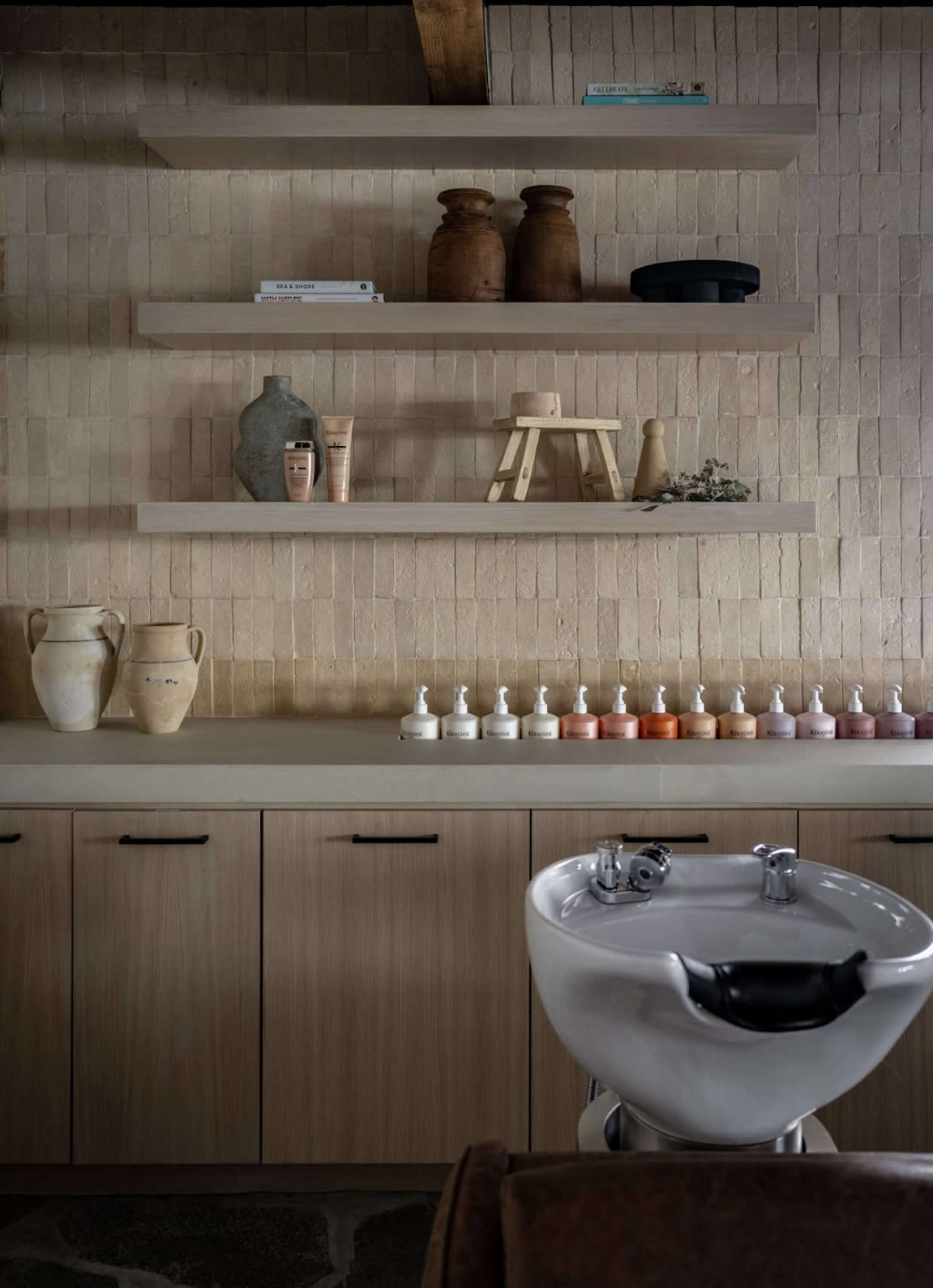 A salon space features a wash basin in the foreground, with neatly arranged shelves displaying various decorative items and skincare products against a textured wall.