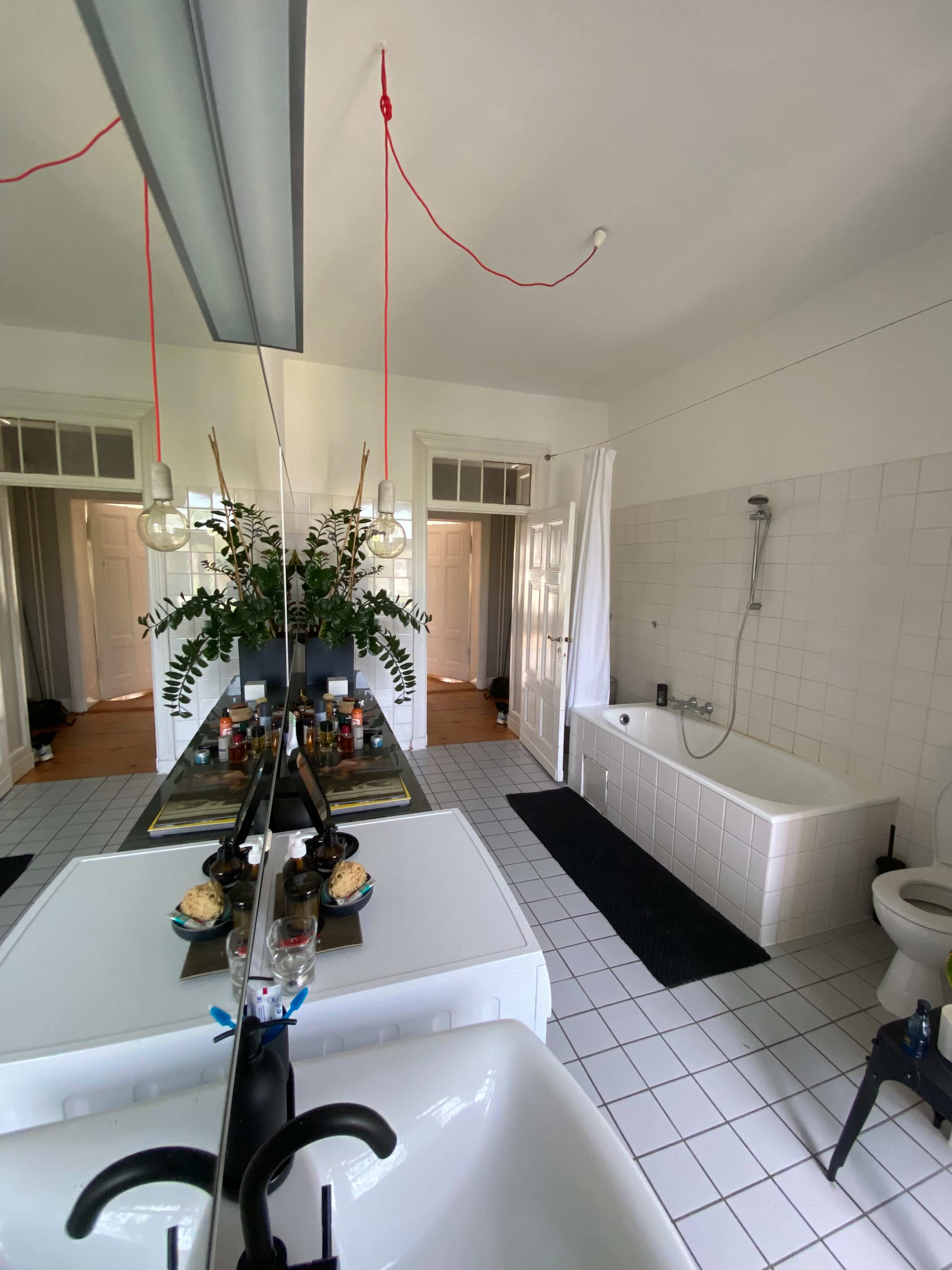 A bathroom features a large mirror, a freestanding bathtub, a toilet, and a sink, with decorative plants on the counter and white tiled flooring.