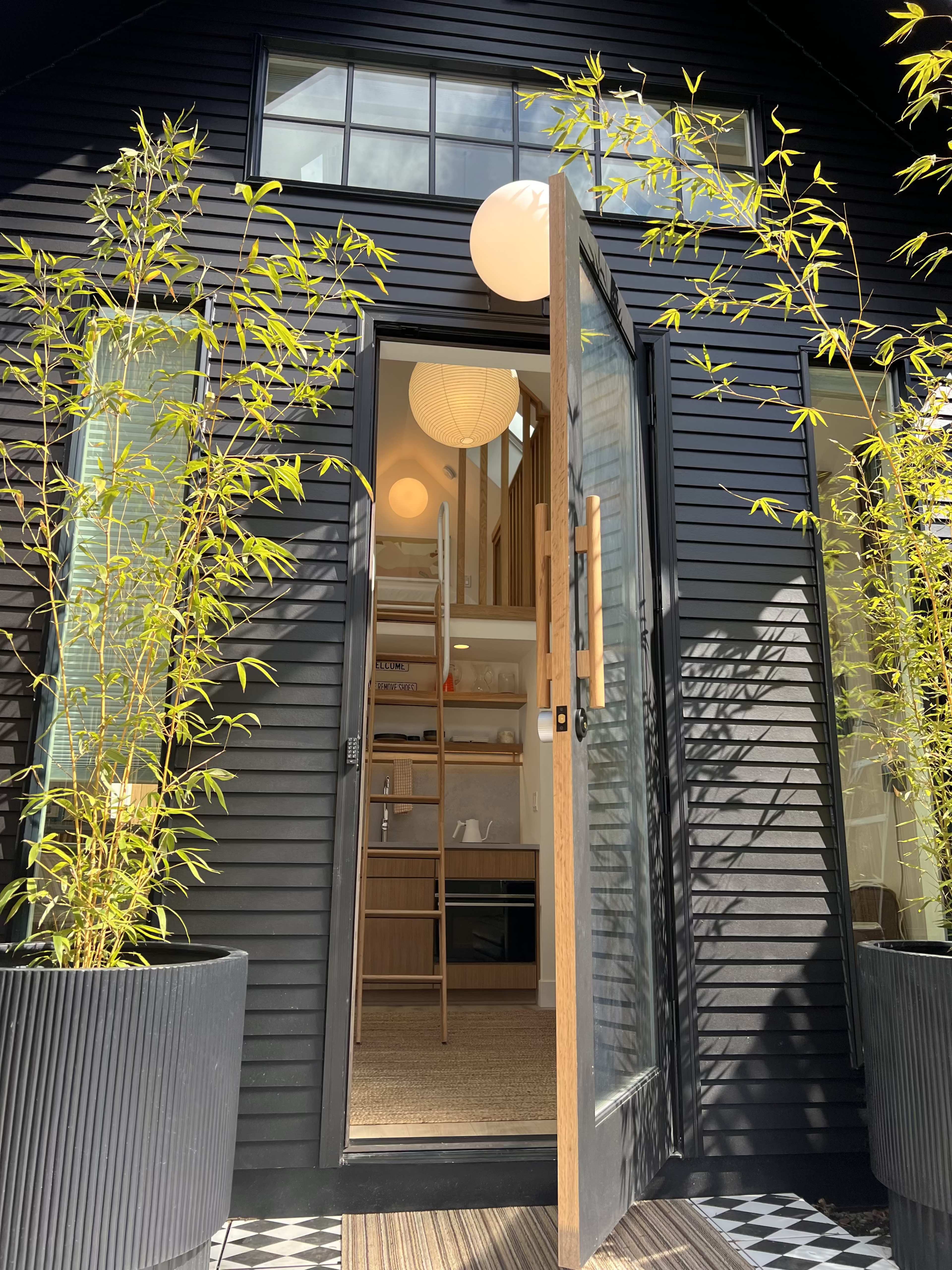 A modern black house features an open front door flanked by tall bamboo plants in planters, leading into a bright interior with a visible ladder and round pendant lights.