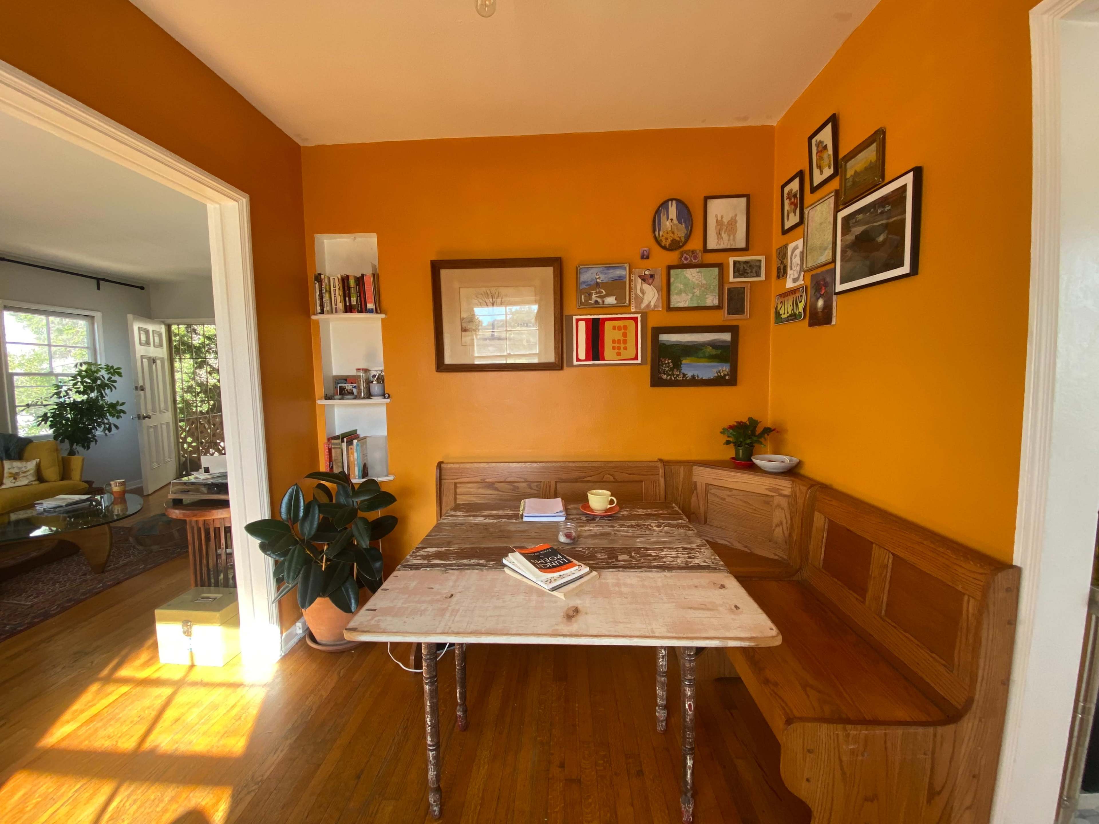 A wooden table with a bench is positioned in a bright orange room adorned with framed pictures and a small bookshelf.