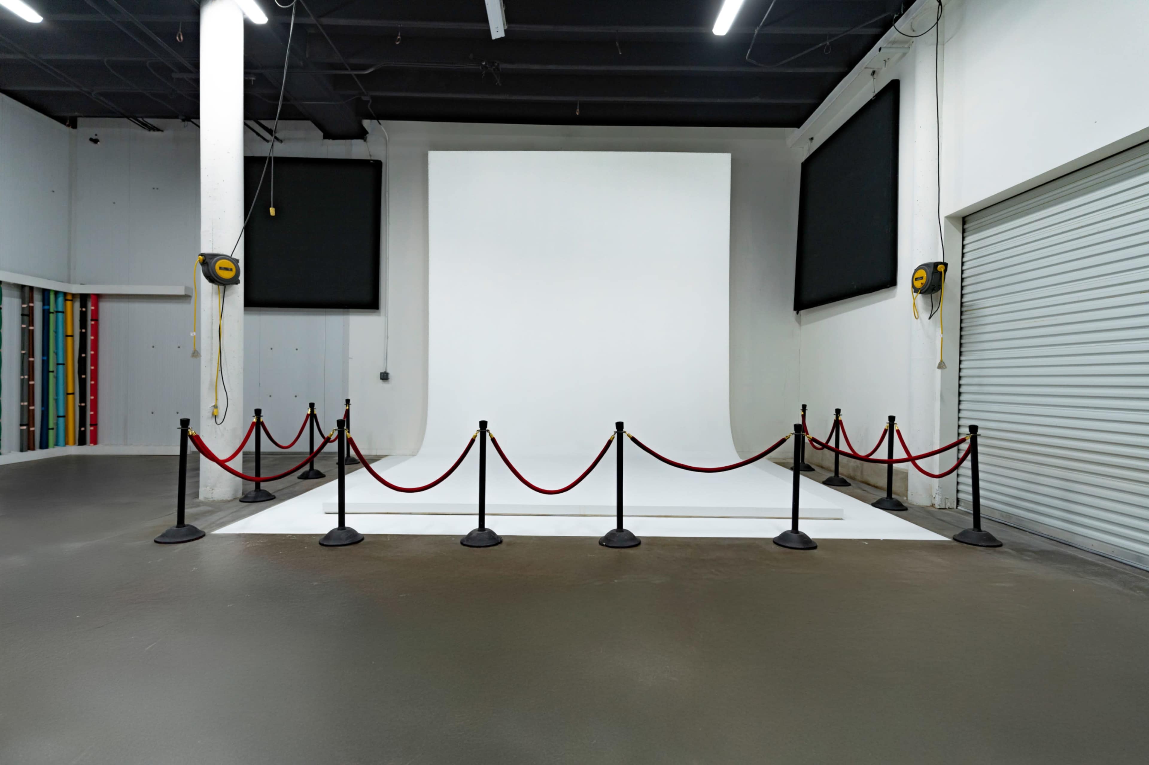 The image shows a spacious, empty studio with a large white backdrop, black posts, and red ropes forming a barrier.